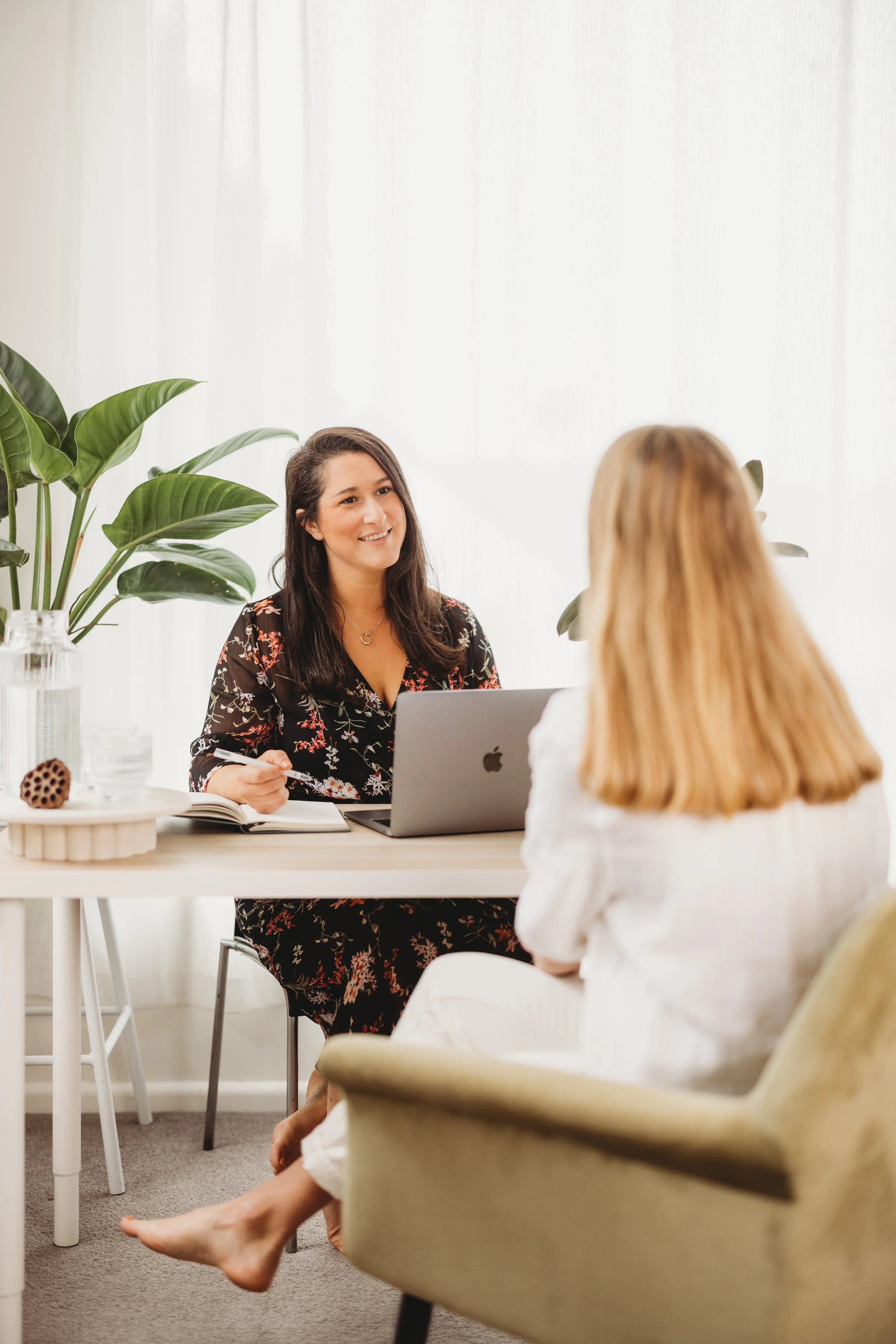 Two women having a conversation in a bright, modern room with large plants. One woman is sitting at a desk with a laptop, and the other woman is seated in an armchair wearing light-colored shoes and a white outfit.