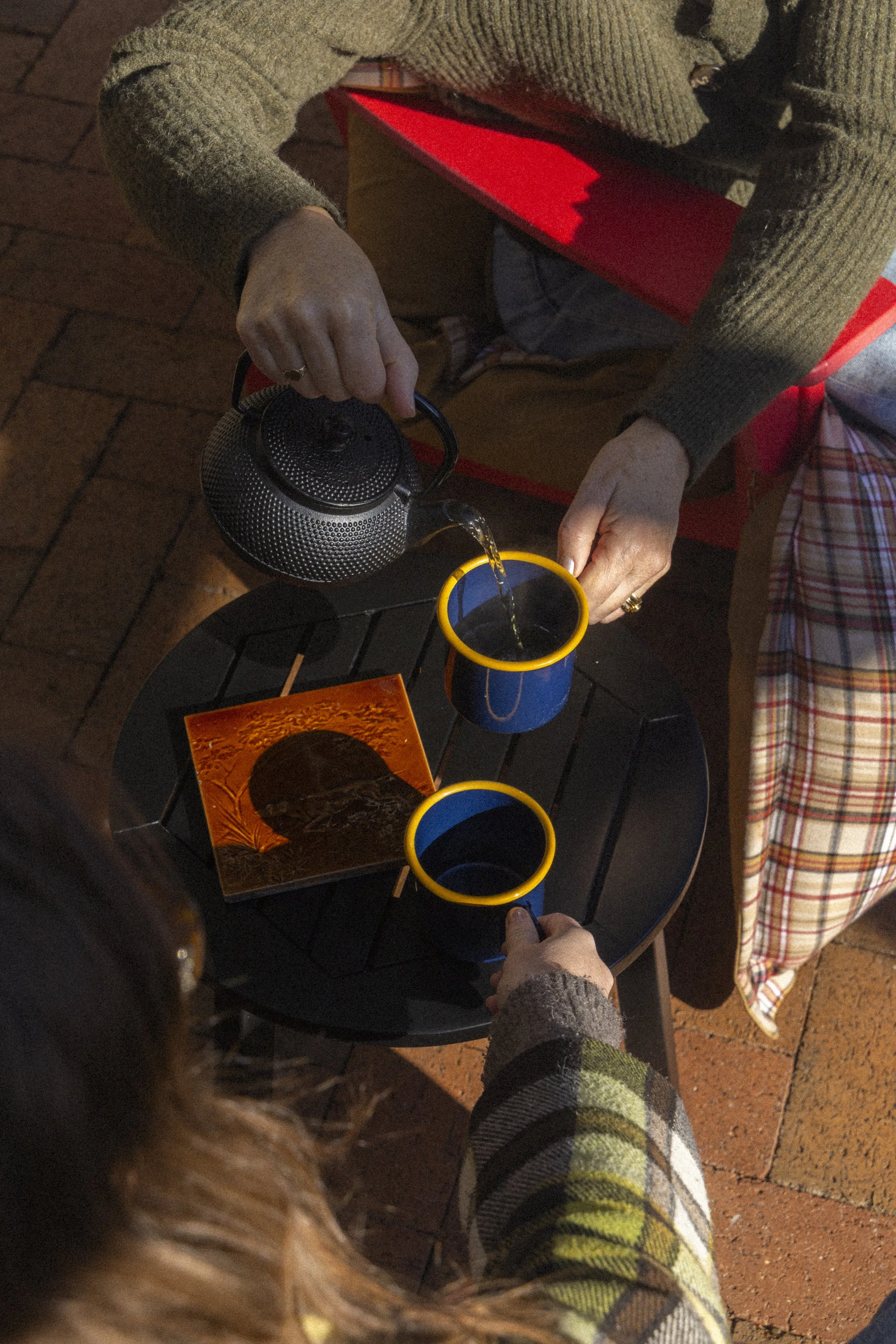 Person pouring hot water from a black metal kettle into a blue mug with a yellow rim on a black outdoor table, with another mug and a book nearby, on a brick surface.