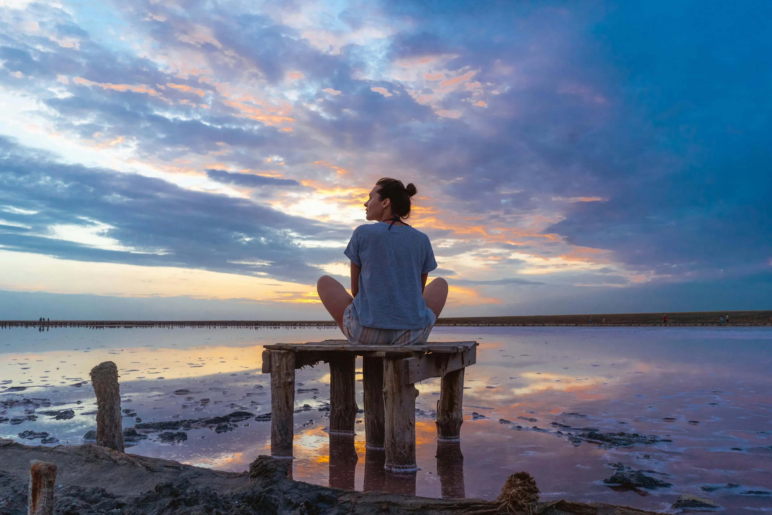 A woman sitting cross-legged on a wooden dock by the water during sunset, with a sky filled with colorful clouds and small people walking in the distance.