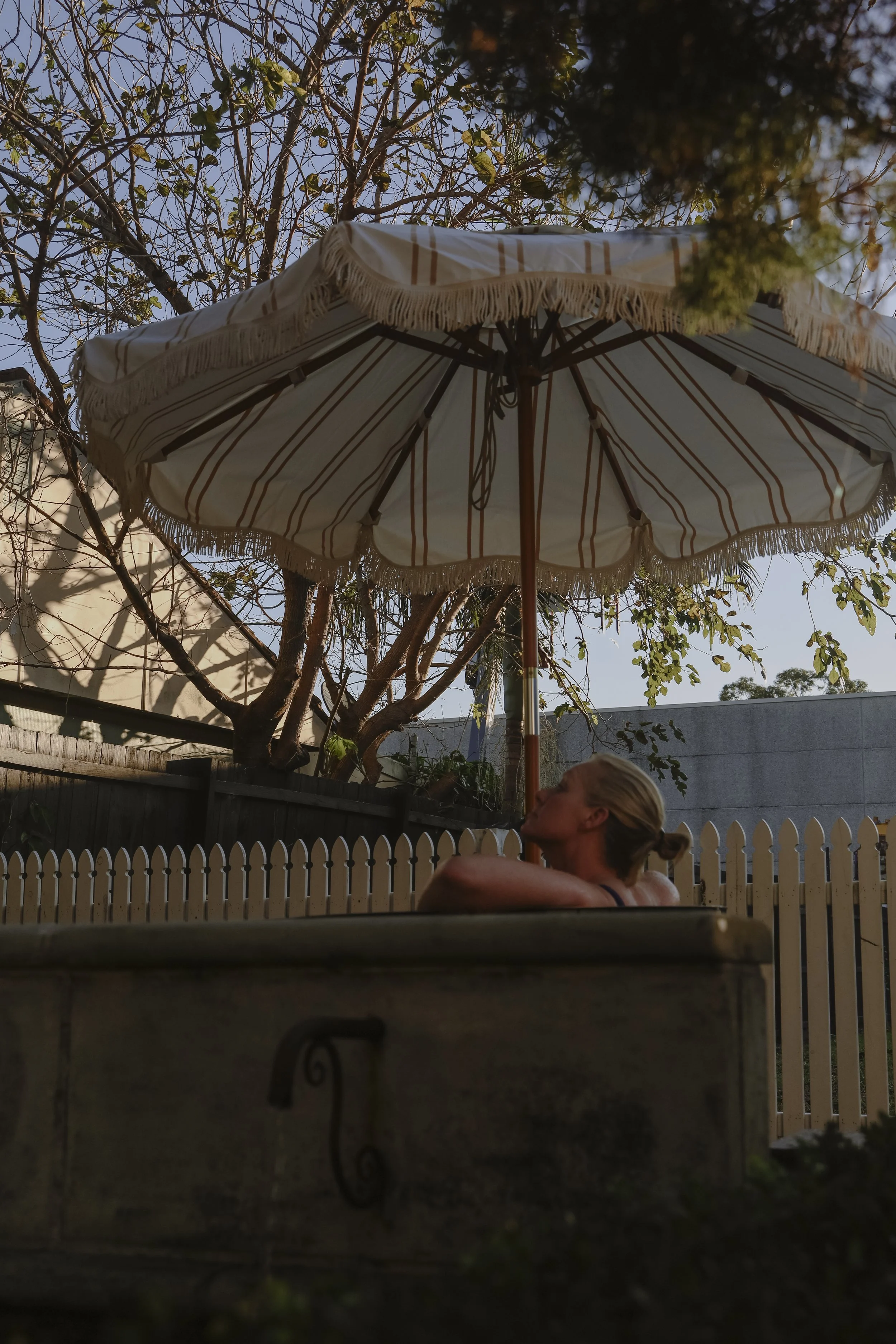 A woman with blonde hair tied in a bun relaxing in an outdoor hot tub, with a striped umbrella providing shade overhead, surrounded by a white picket fence, trees, and a blue sky in the background.