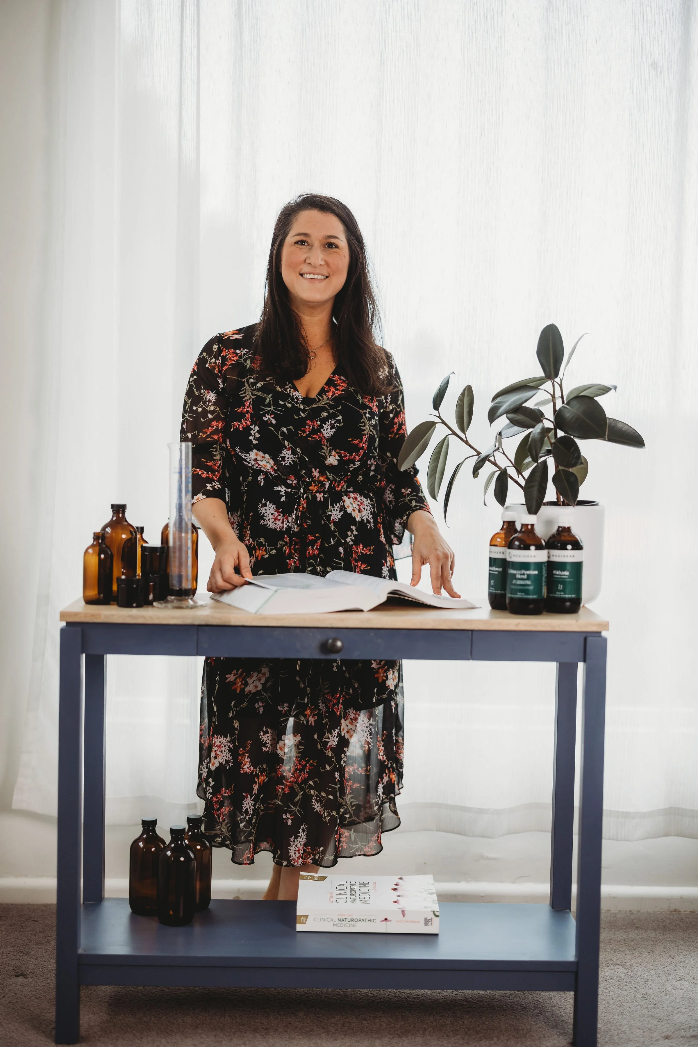 A woman in a black floral dress standing at a table with brown bottles, a white book titled 'Clinical Naturopathic Medicine,' and potted plant. The background has white curtains.