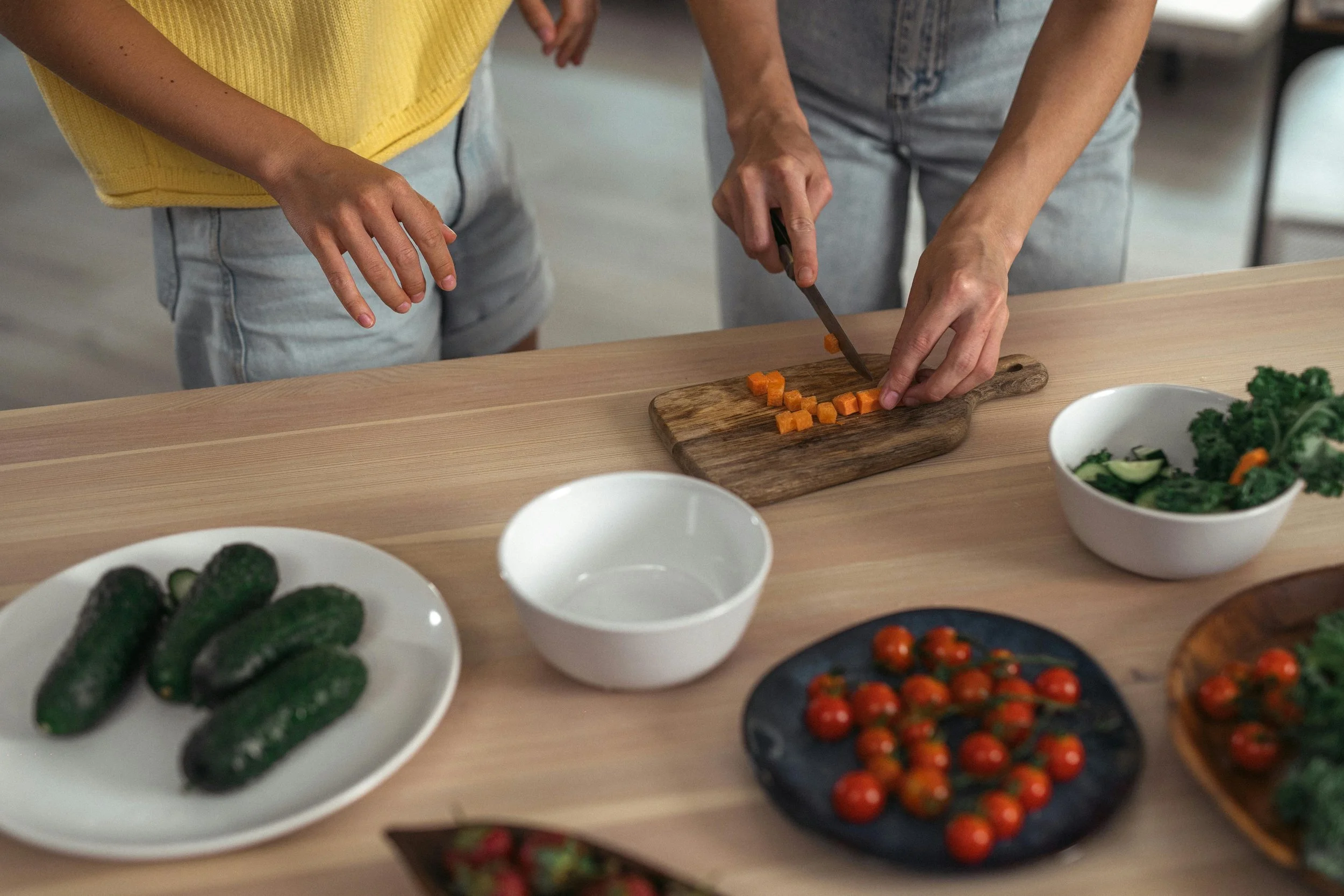 People chopping carrots on a wooden cutting board alongside various bowls of salad ingredients, including cherry tomatoes, cucumbers, and greens, on a table.