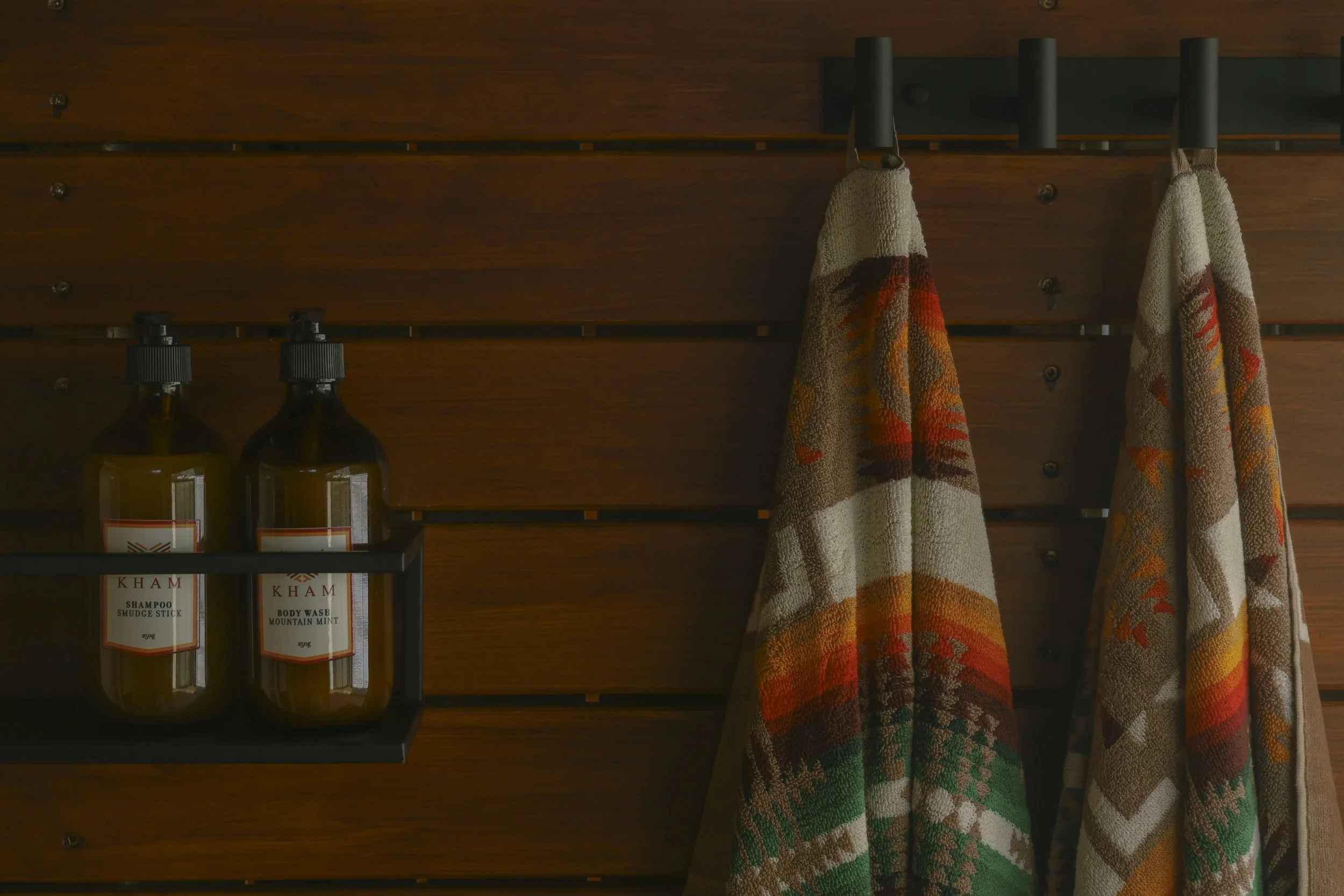 Wooden wall with two bottles of shampoo and body wash, and two towels with colorful patterns hanging on hooks.