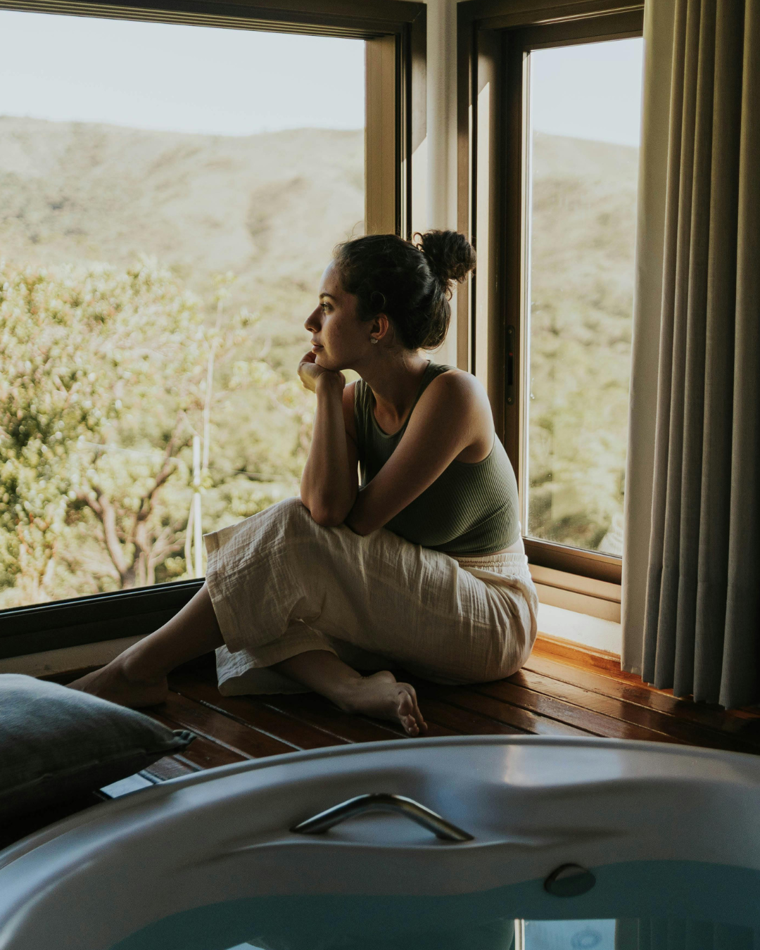 A woman sitting on a wooden windowsill, looking outside at a green landscape, resting her chin on her hand.