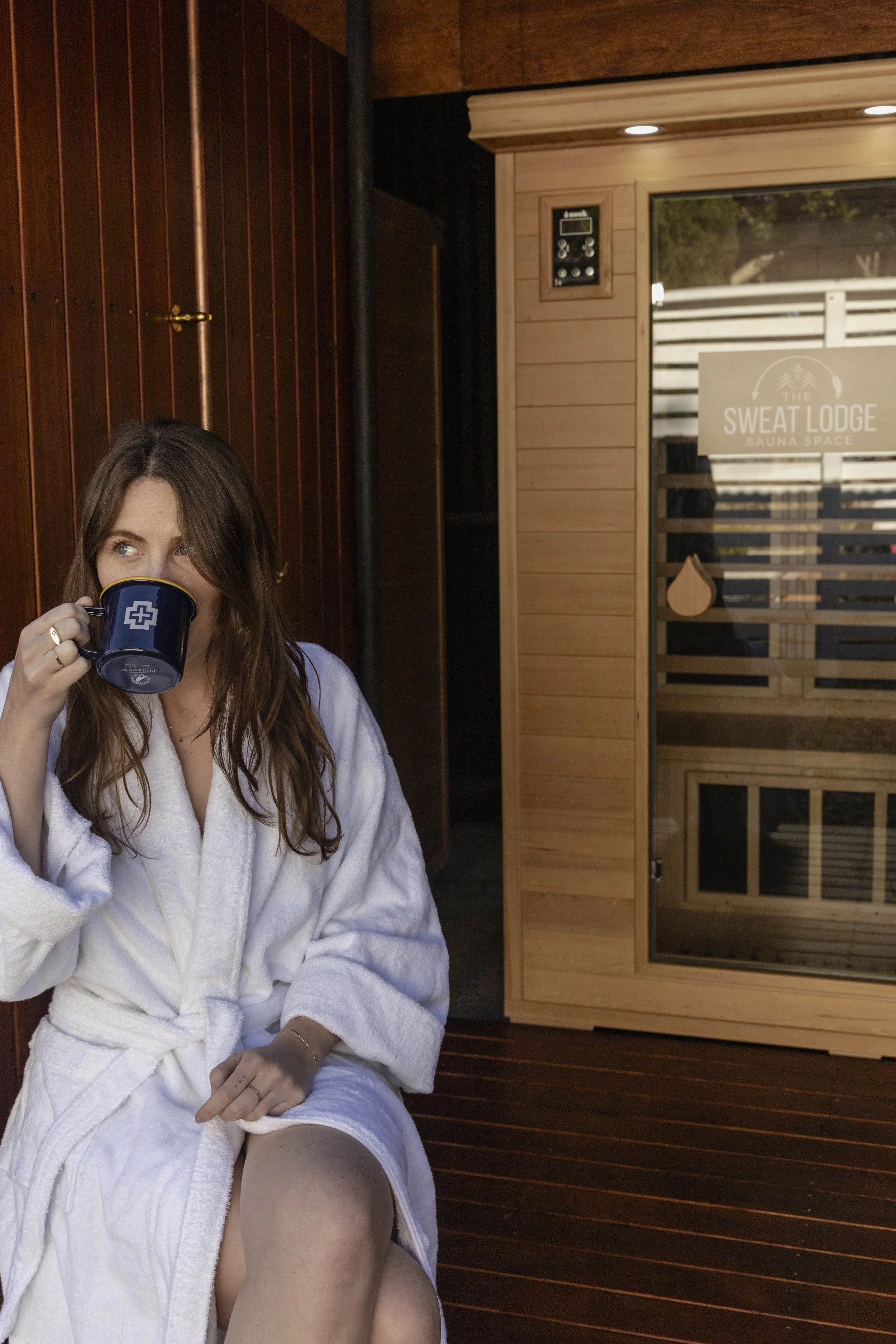 A woman with brown hair and fair skin, wearing a white bathrobe, sitting in a sauna room drinking from a dark blue mug with a white cross logo, with a sauna room sign visible in the background.
