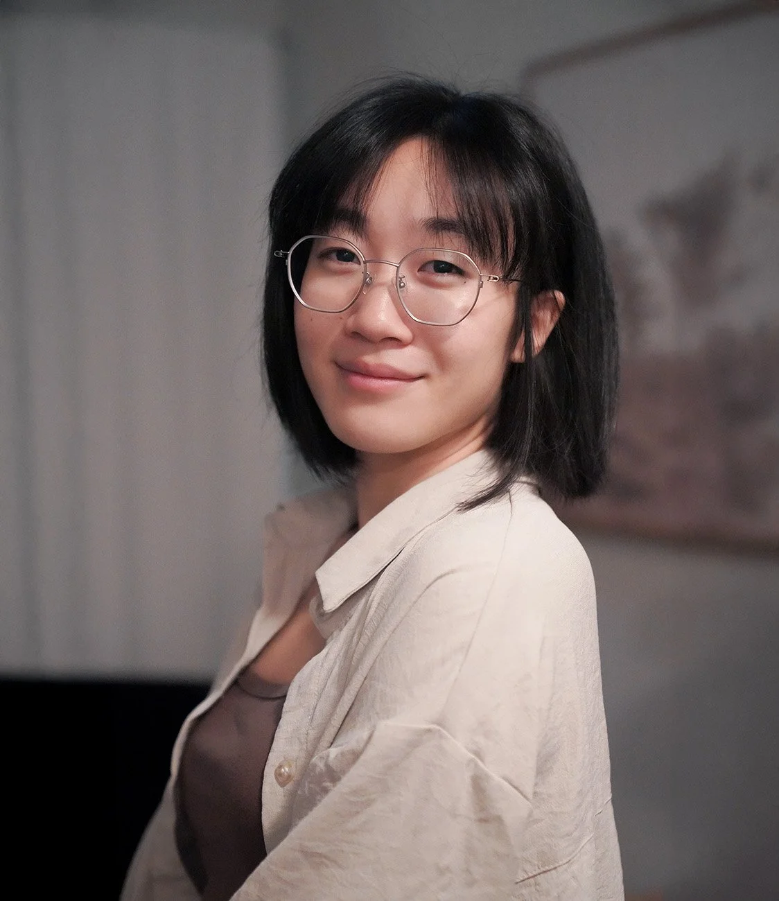 A young woman with short black hair and glasses, smiling while looking at the camera, wearing a beige shirt.