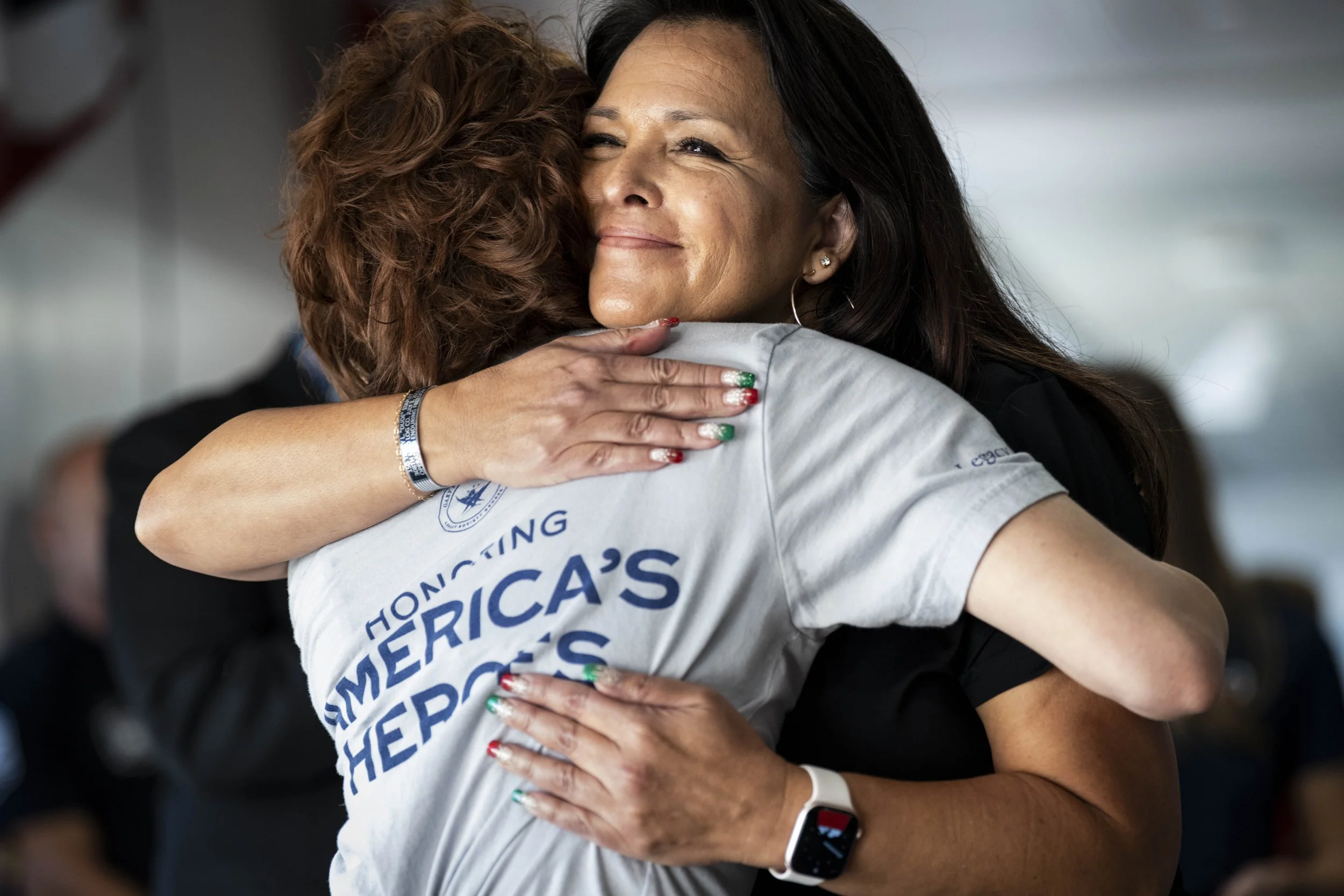 Two women hugging, one wearing a white shirt with blue text, and the other wearing a black outfit. The woman in black is smiling and has her eyes closed.