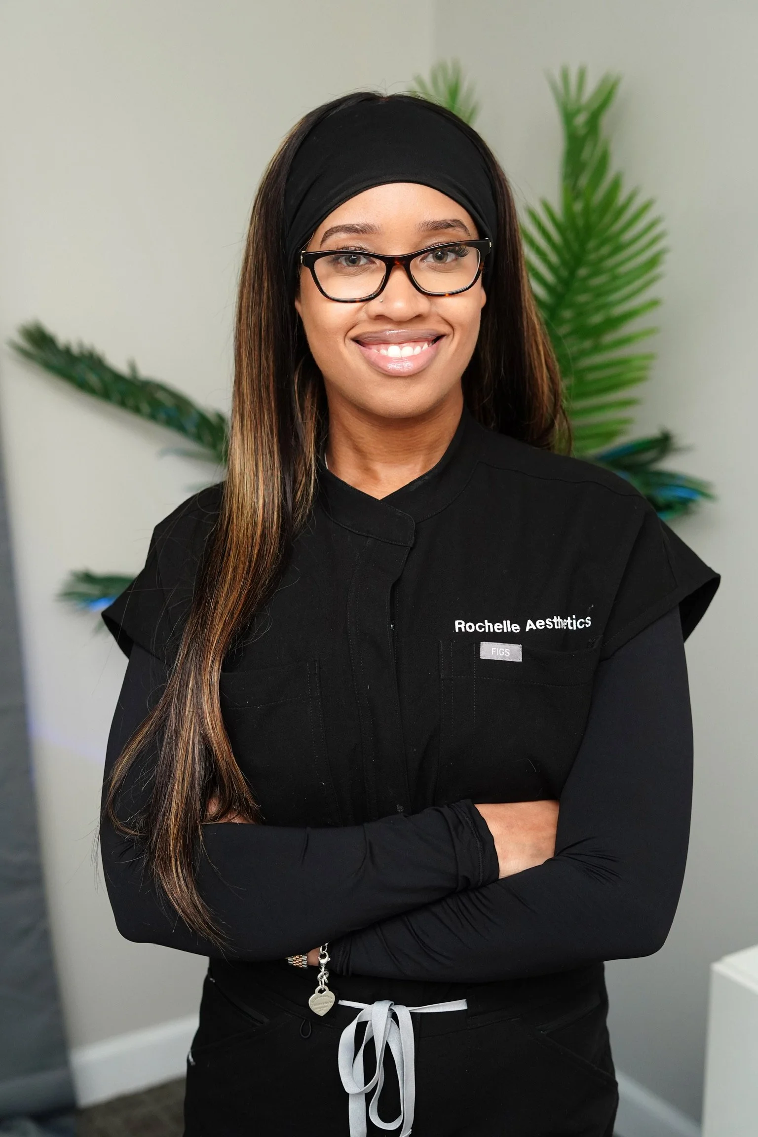 A woman with short platinum blonde hair wearing glasses, a black scrub top, a gold necklace, and a colorful floral tattoo on her forearm, smiling against a dark blue background.