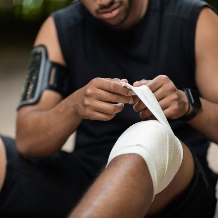 Man wrapping a bandage around a bent knee while seated outdoors.