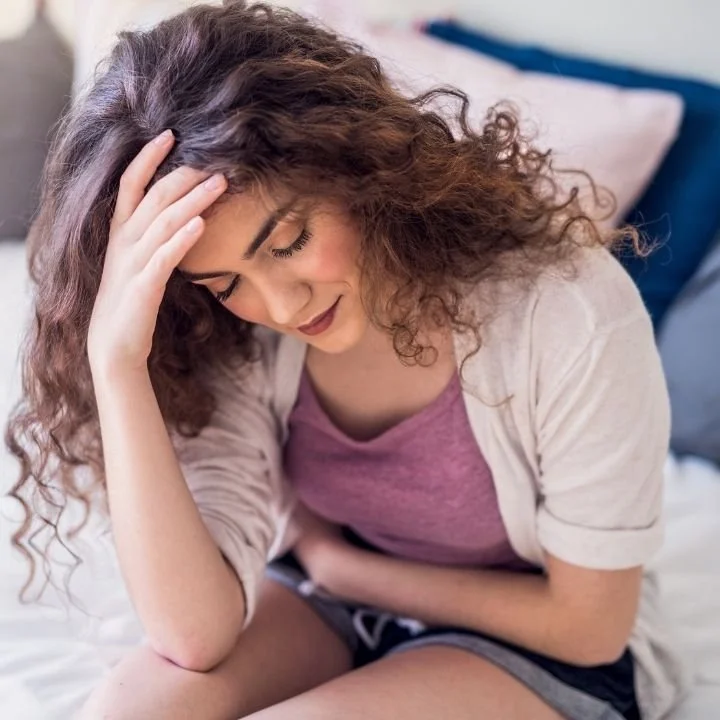 Woman sitting on bed holding head and abdomen while leaning forward.
