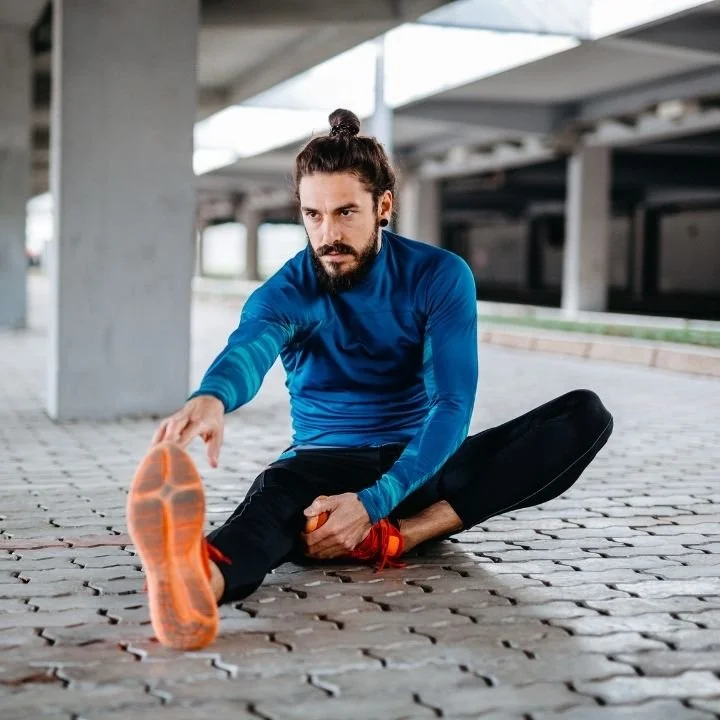 Adult stretching one leg forward while seated on a paved surface outdoors.