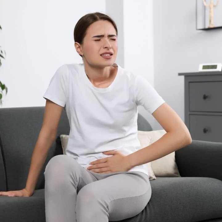 Woman seated on couch holding lower abdomen with visible discomfort.