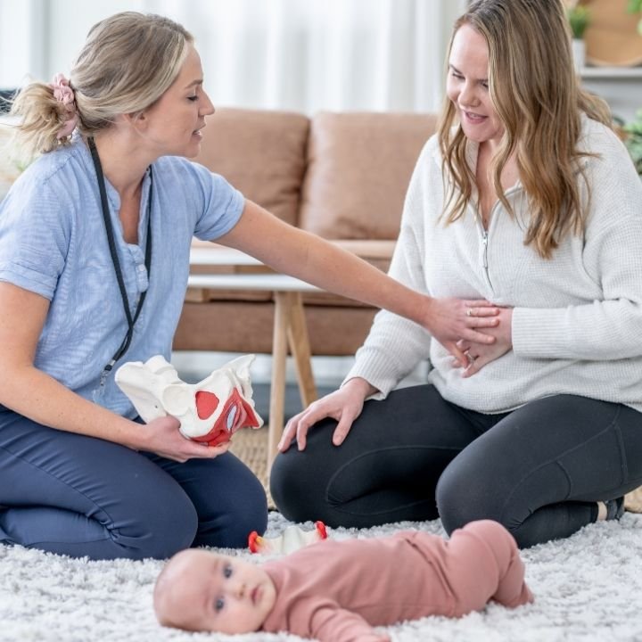 Pelvic floor therapist discussing recovery with mother while baby lies on mat.