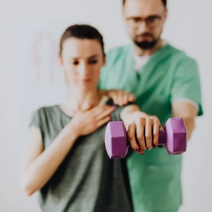 Therapist supervising patient performing light dumbbell shoulder raise during rotator cuff rehab.