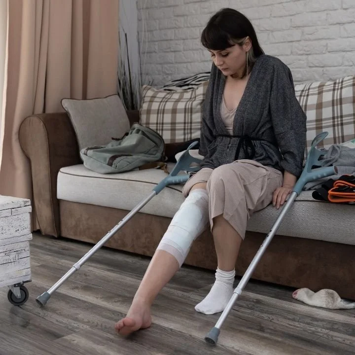 Woman sitting on a couch with crutches and a wrapped knee resting on the floor.