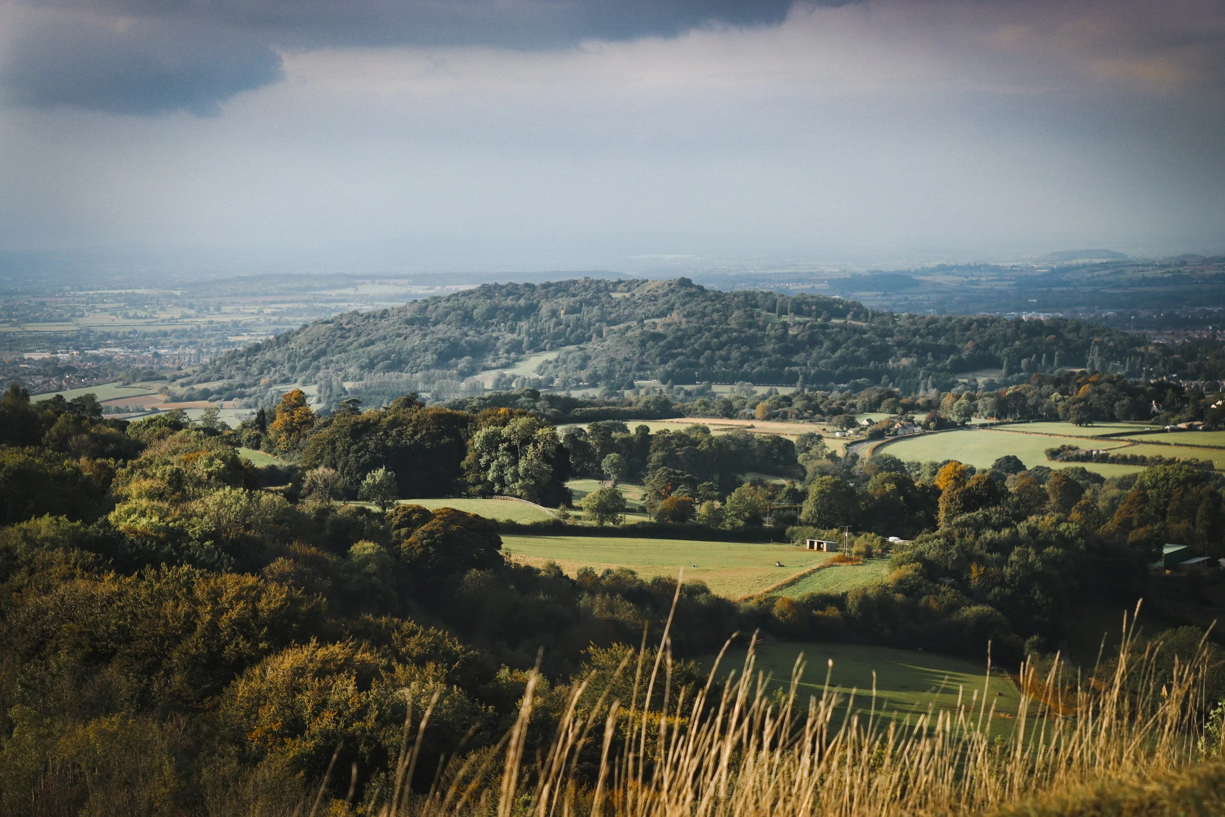 Rolling Cotswold countryside landscape with fields, hills and trees under a cloudy sky.