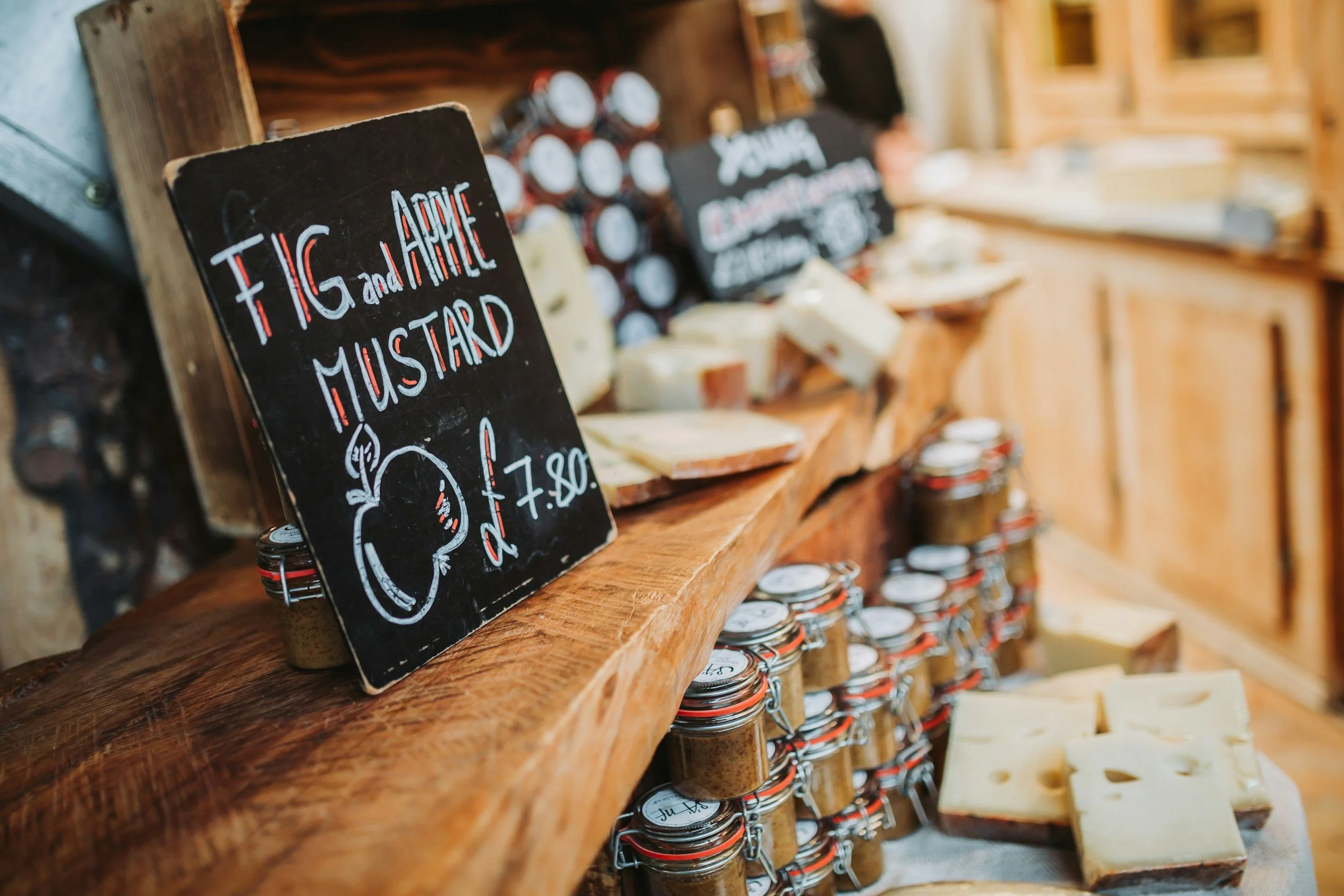 Artisan cheese and condiment stall with jars, cheese blocks and handwritten chalkboard sign.