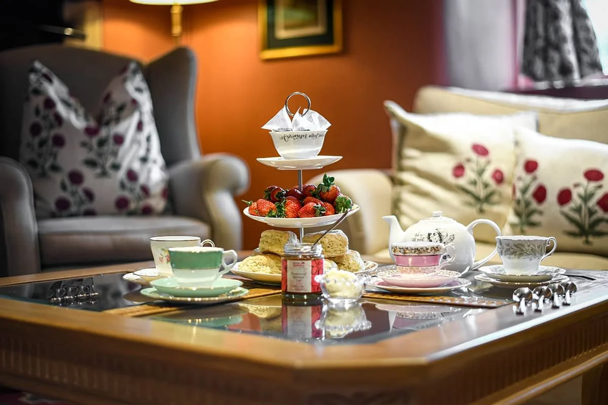Afternoon tea setup indoors with tiered stand, scones, strawberries, teapot and teacups on a coffee table.