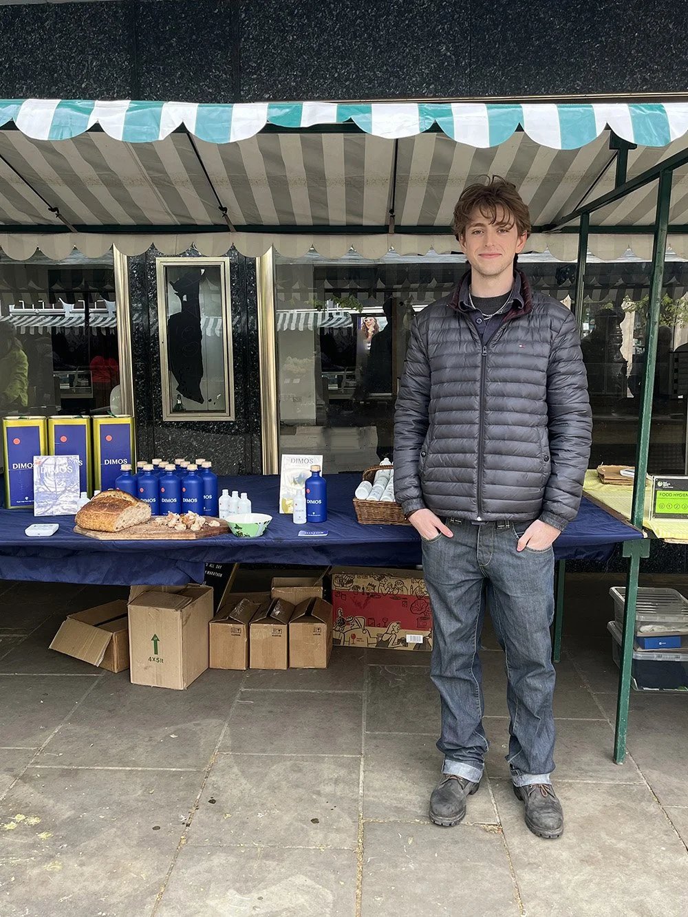 Young vendor standing behind a DIMOS olive oil stall with bottles and food display.