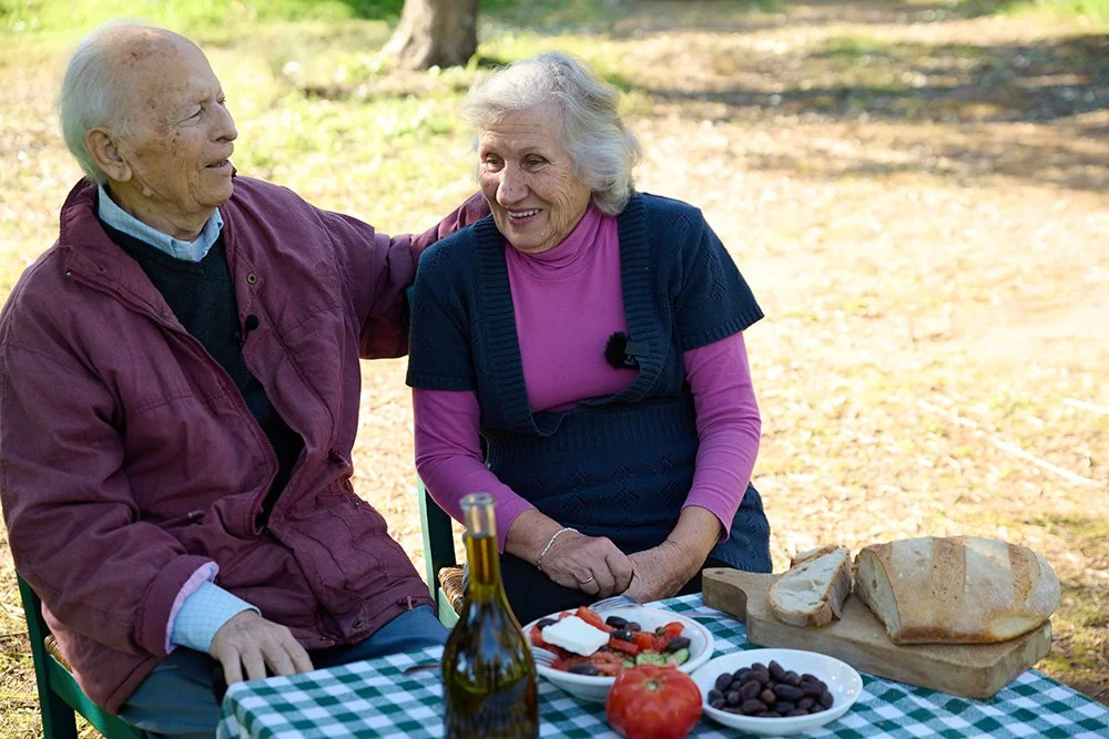 Elderly couple sitting together outdoors in an olive grove.
