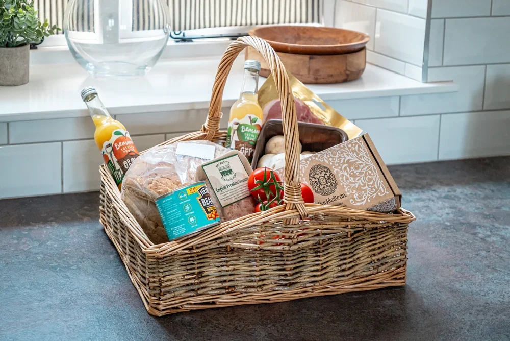 Cotswold breakfast hamper in a wicker basket with bread, eggs, bacon, juice and local produce on a kitchen floor.