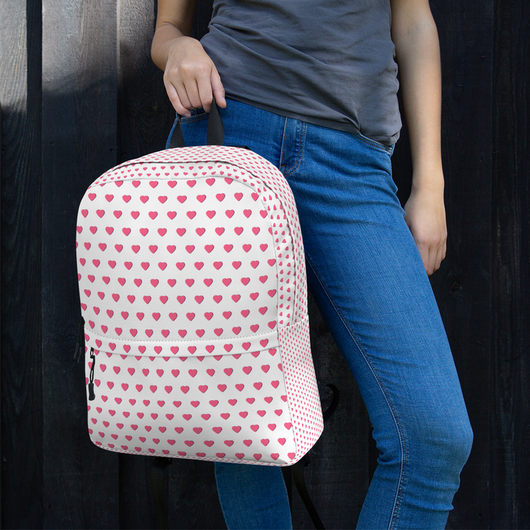 Person standing outdoors holding a white backpack with pink heart patterns, wearing blue jeans and a gray T-shirt.