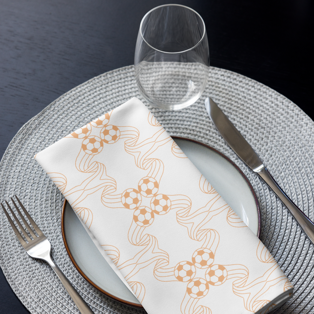 A table setting with a white napkin featuring a soccer ball pattern, a wine glass, a white plate, a fork, and a knife on a woven gray placemat on a dark table.
