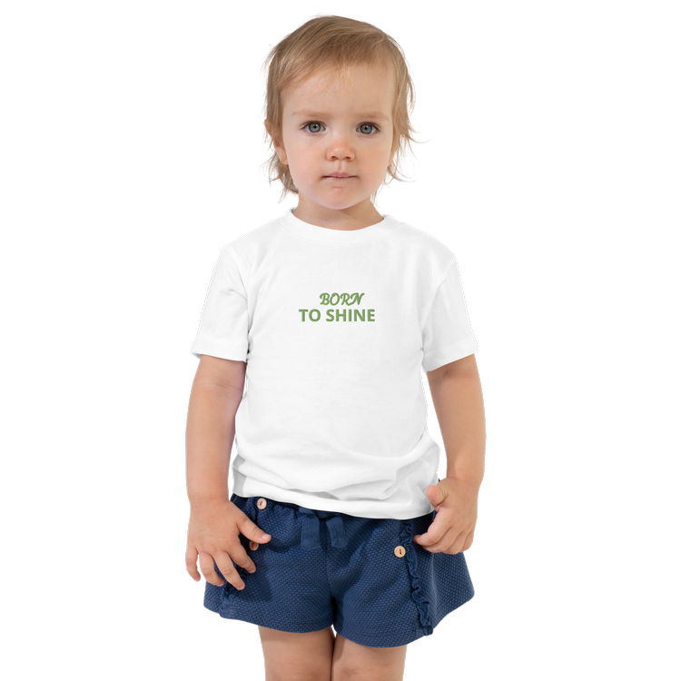 Young child wearing a white t-shirt with the text 'Born to Shine' in green letters, paired with blue shorts. Custom Apparel