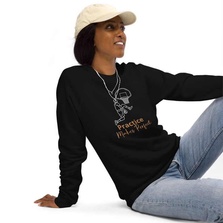 A person wearing a beige cap and a black sweatshirt with a basketball player graphic and the text "Practice Makes Perfect." They are sitting with one knee bent, wearing light blue jeans. Unique Motto Design on Casual Wear.