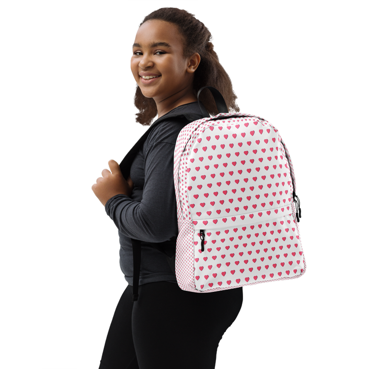 A smiling young girl with curly hair holding a black strap backpack over her shoulder, wearing a black long sleeve shirt and black pants, with a white backpack decorated with pink hearts.