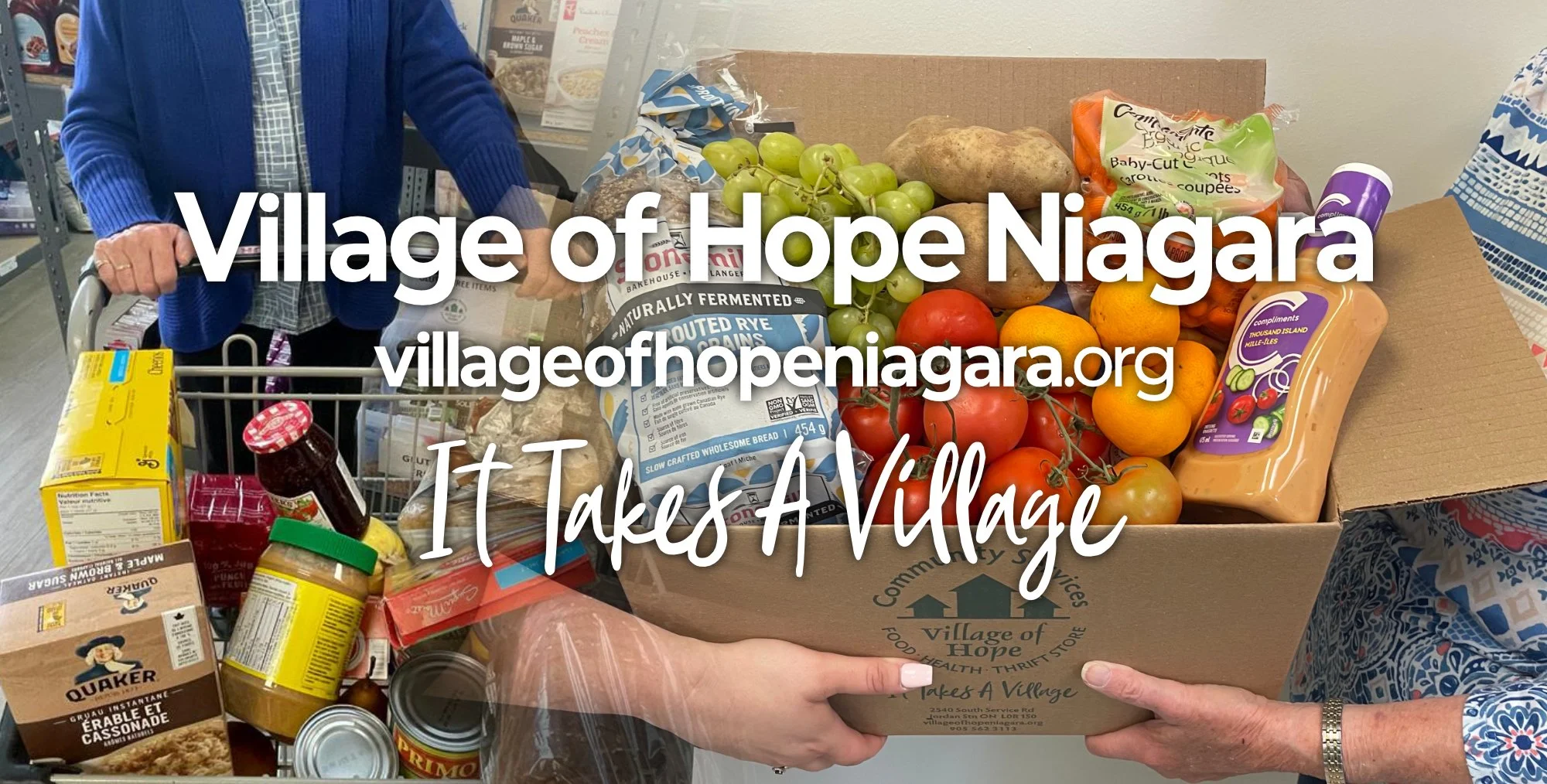 A person holding a cardboard box filled with fresh vegetables and groceries at a food bank or thrift store. Items include tomatoes, oranges, grapes, potatoes, juice, bread, and canned goods. The box has the logo and contact info for Village of Hope.