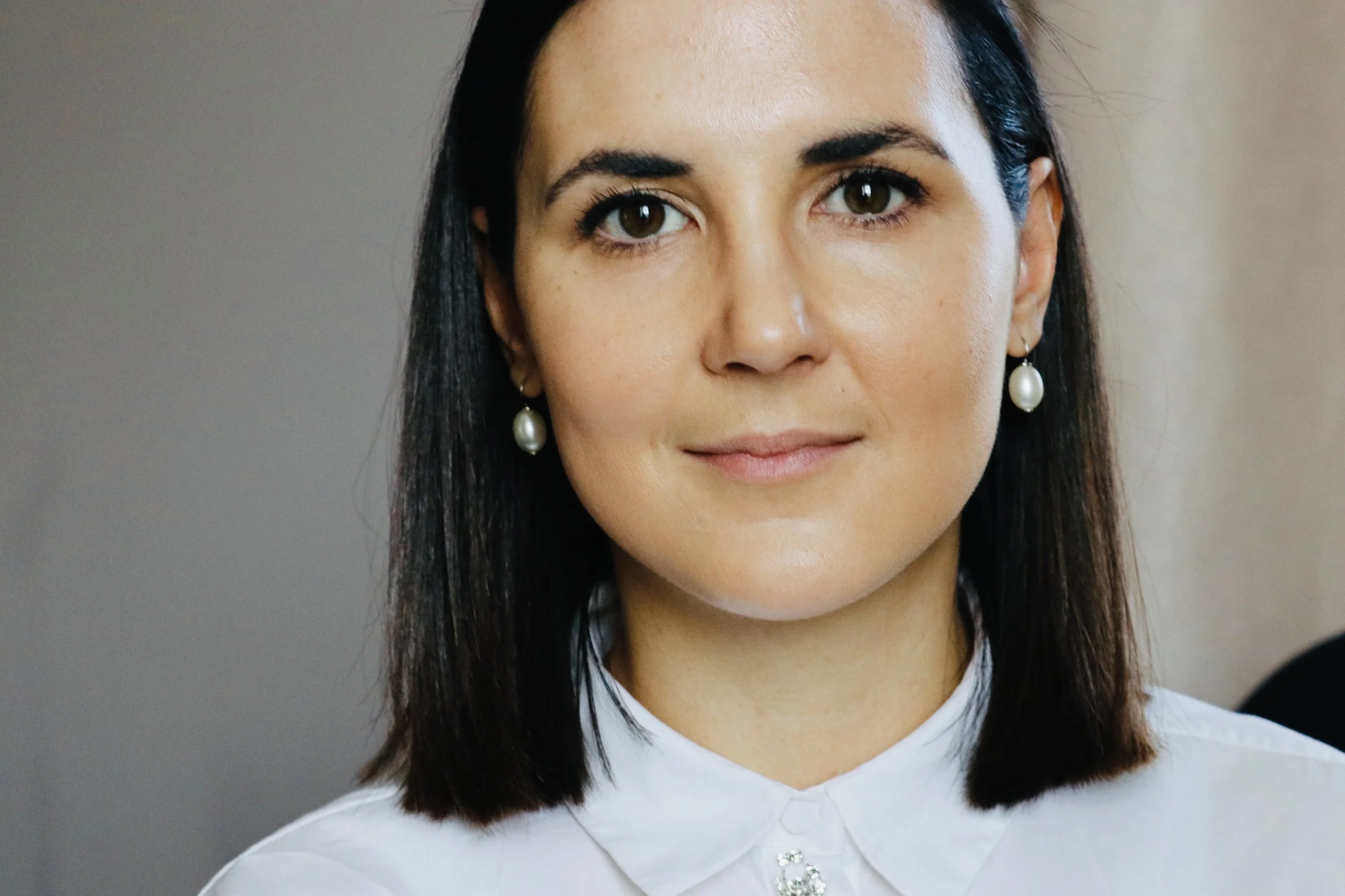 Nahaufnahme einer Frau mit schwarzen Haaren, dunklen Augen, ohrringen mit Perlen, weißem Hemd mit Brosche, lächelnd vor neutralem Hintergrund.