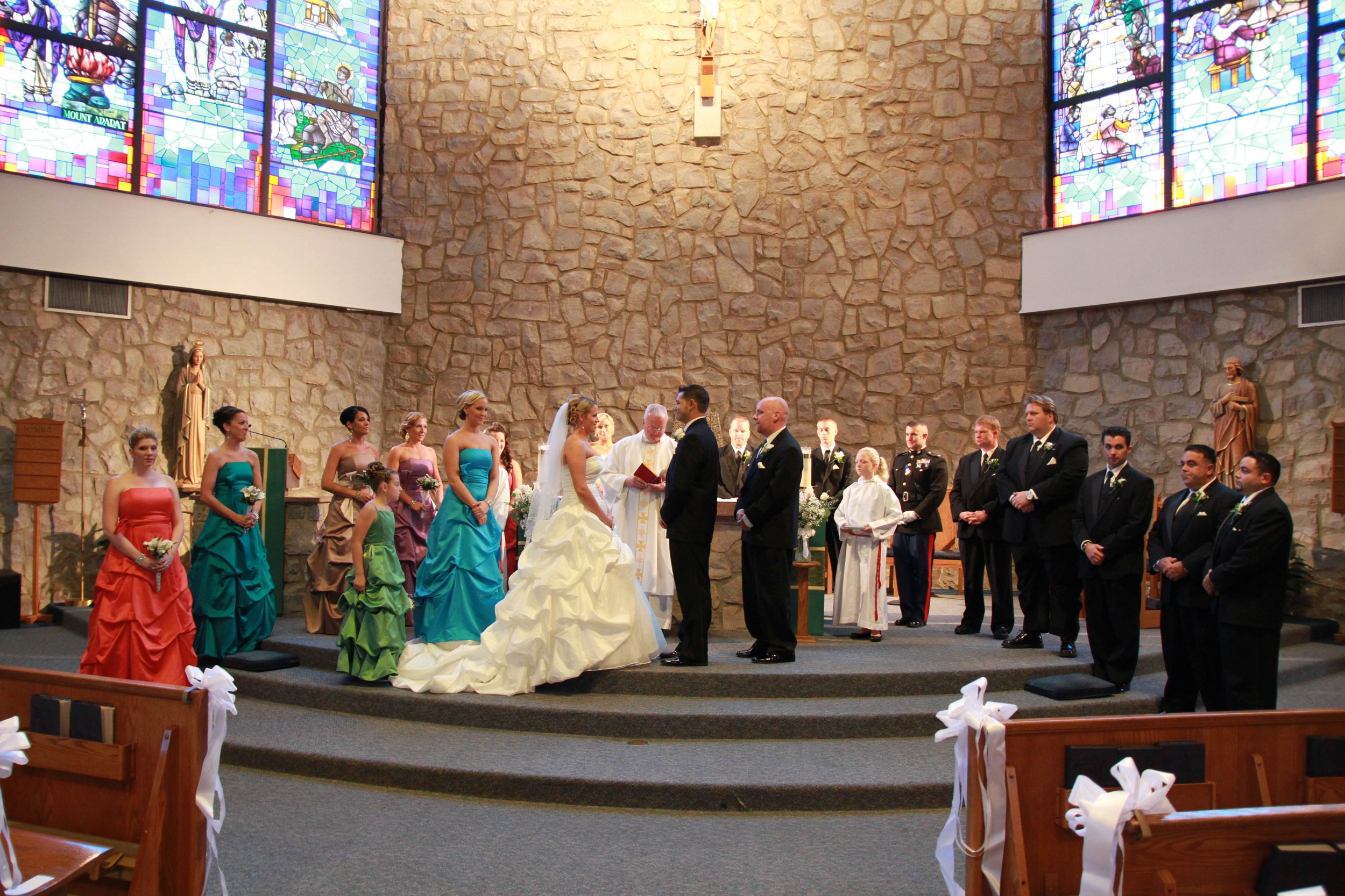 A wedding ceremony inside a church with stone walls and stained glass windows. The bride in a white gown stands at the altar with the groom in a black suit. Bridesmaids in colorful dresses and groomsmen in black suits stand on either side. A priest i