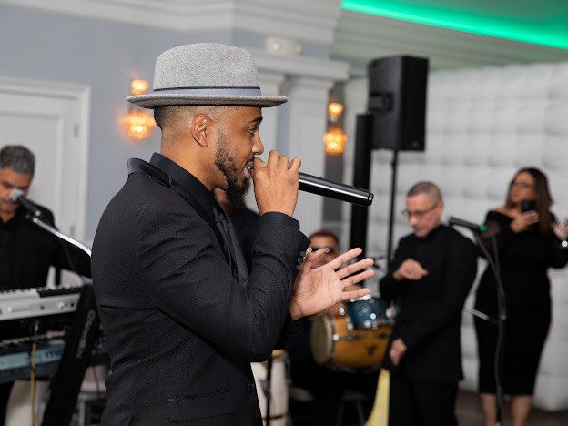 Man in a gray hat singing into a microphone at a live performance with band members in the background.