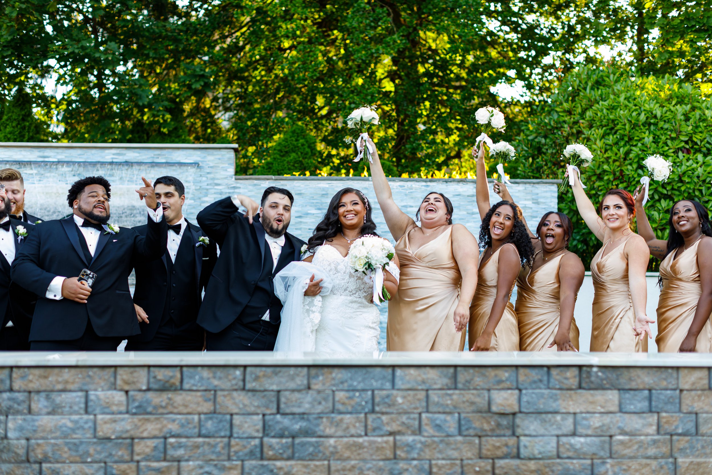 Wedding party group photo with bridesmaids and groomsmen in formal attire, smiling and celebrating outdoors.