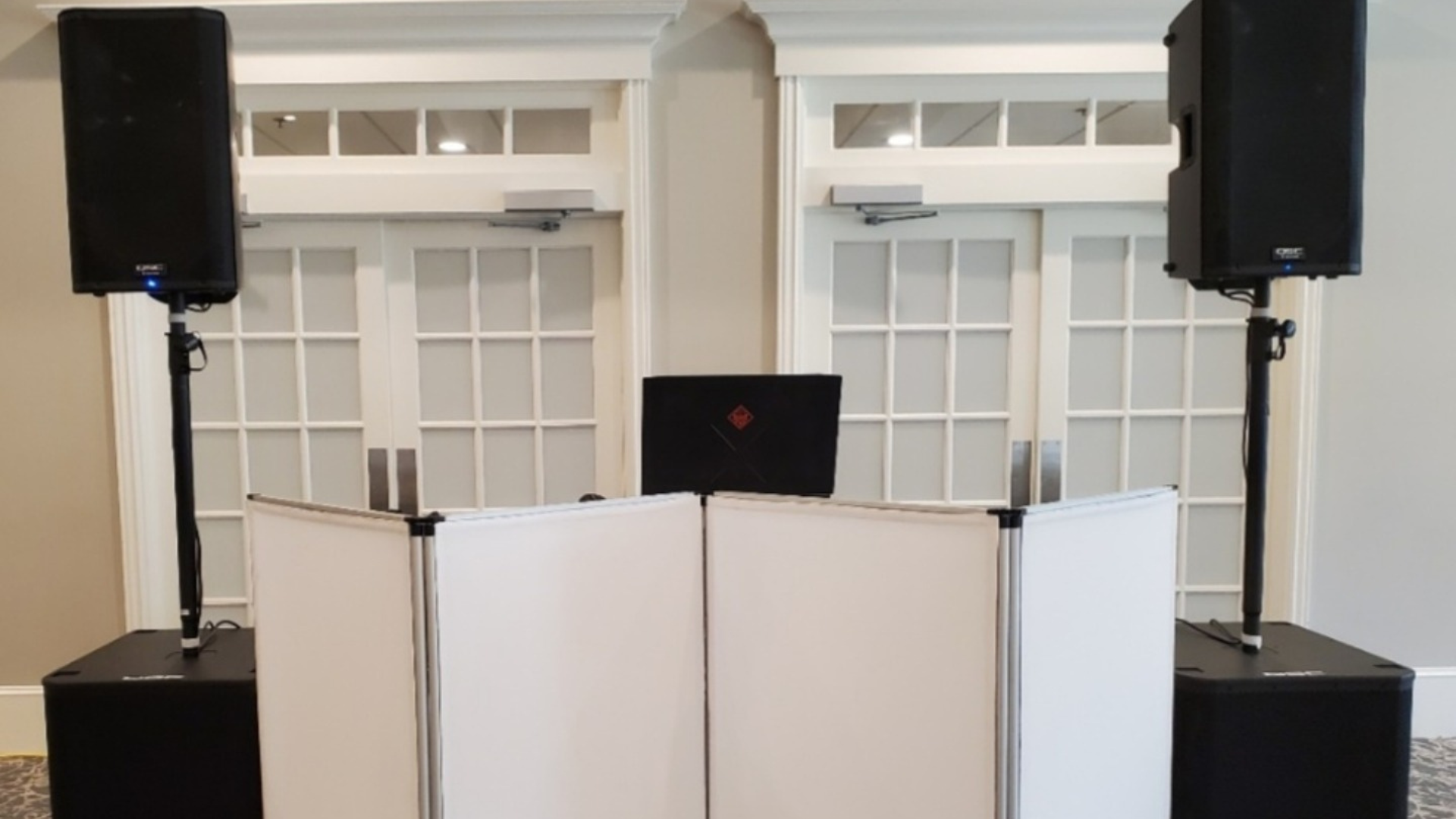 DJ setup with speakers and a laptop behind a white screen in front of double doors.