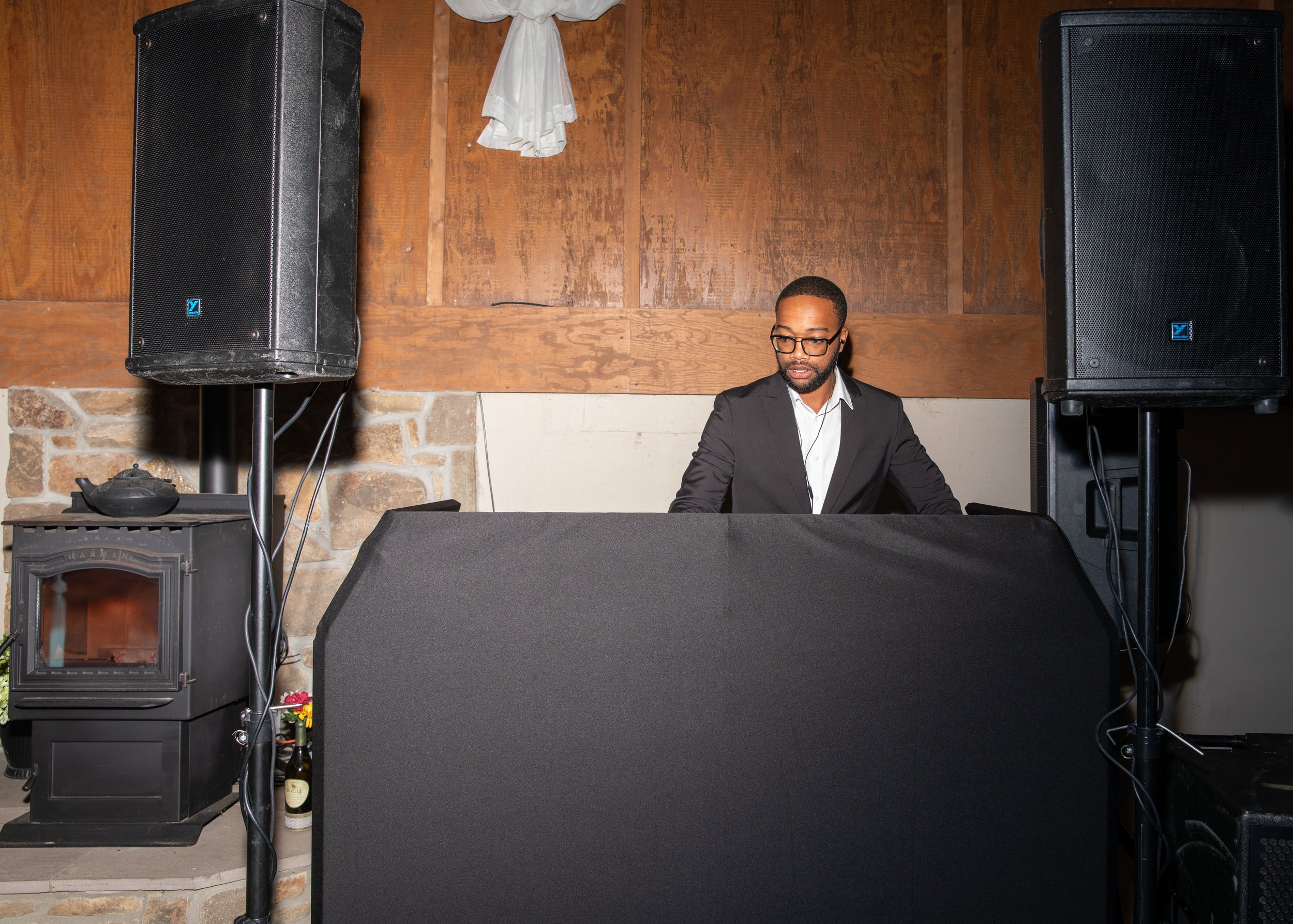 DJ stands behind a covered table with speakers and equipment at an event.
