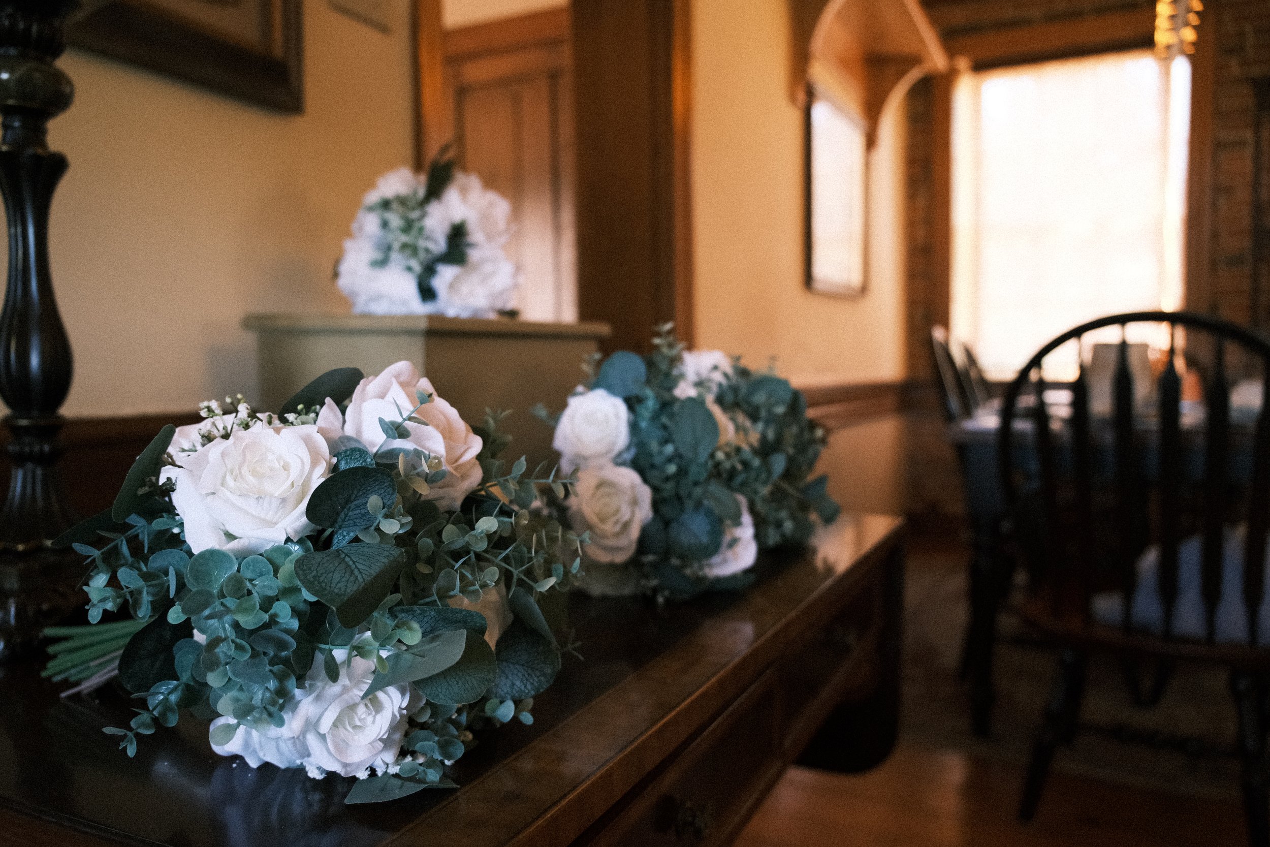Wedding bouquets with white roses and greenery on a wooden table in a warmly lit room.