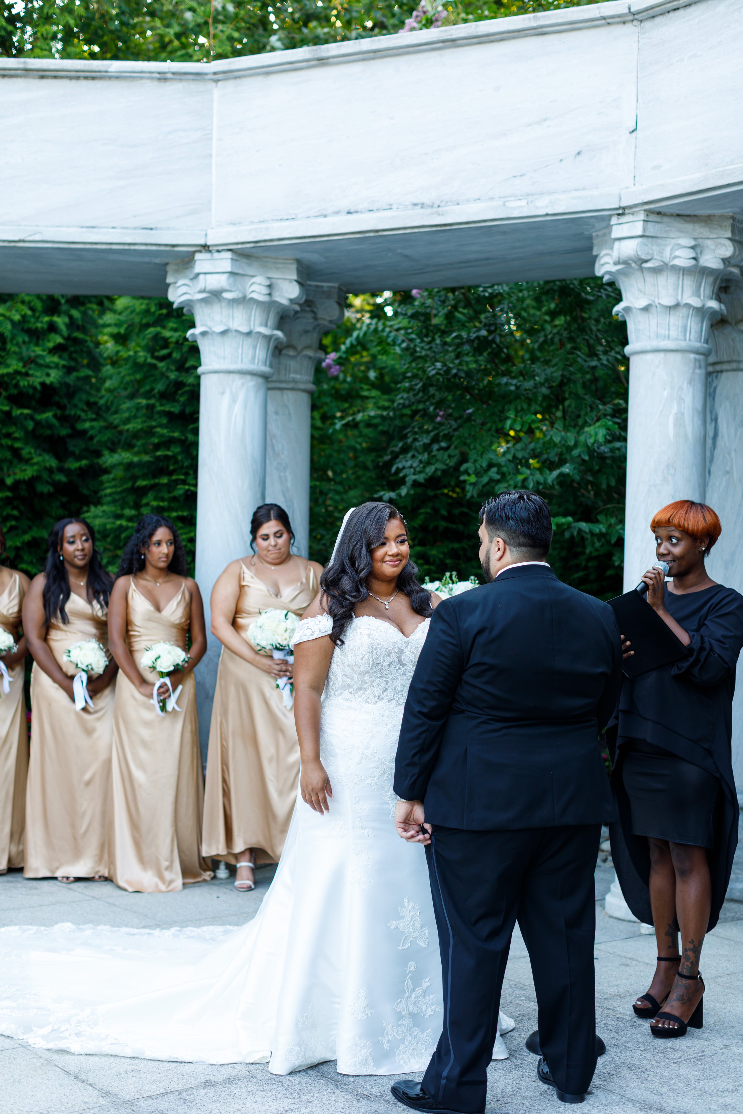 A wedding ceremony taking place outdoors, featuring a bride in a white gown and a groom in a black suit standing under a stone gazebo. Bridesmaids in gold dresses hold bouquets nearby. The officiant is speaking to the couple.
