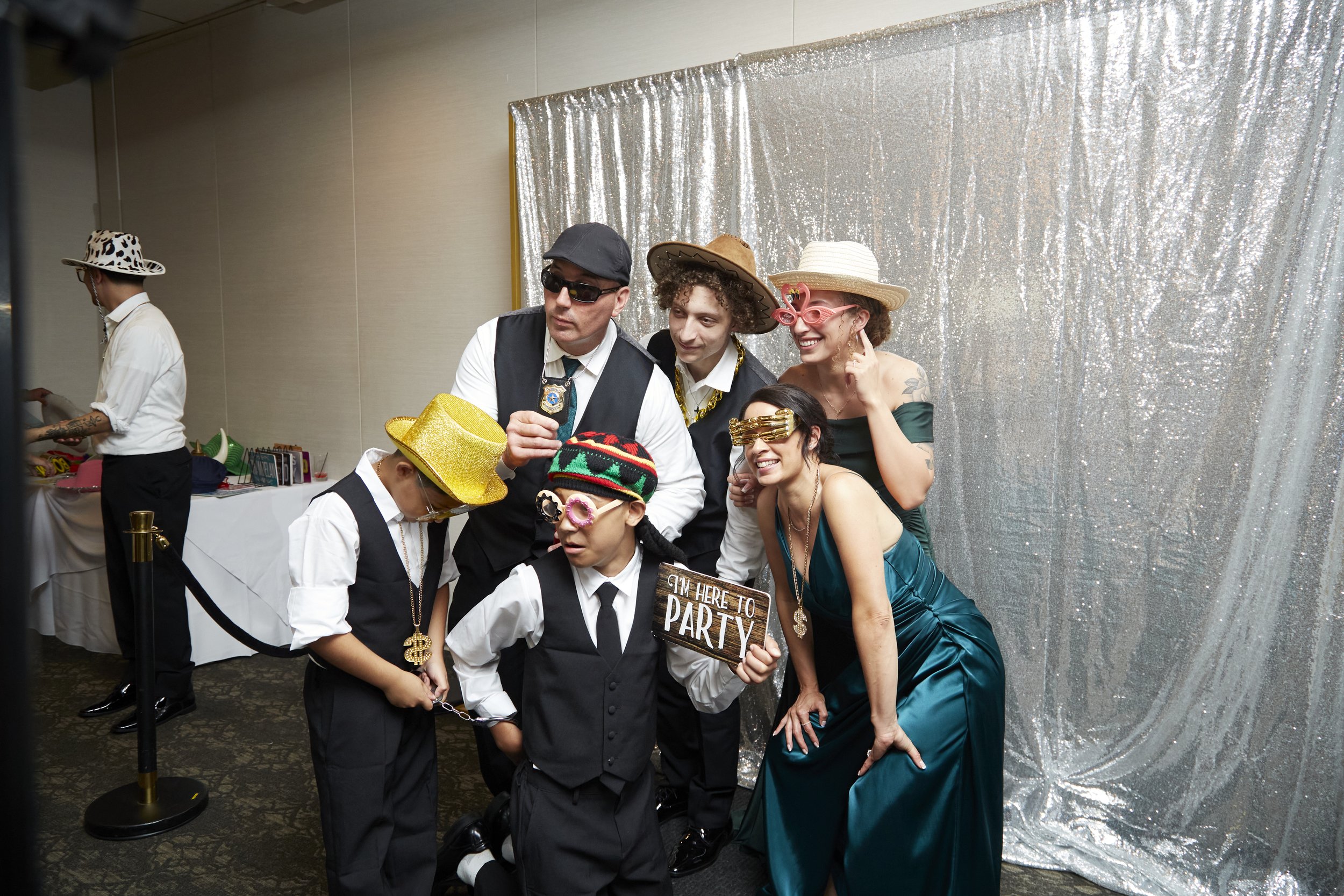 Group of people dressed in funny costumes with hats and sunglasses posing in front of a silver sequin backdrop at a party.