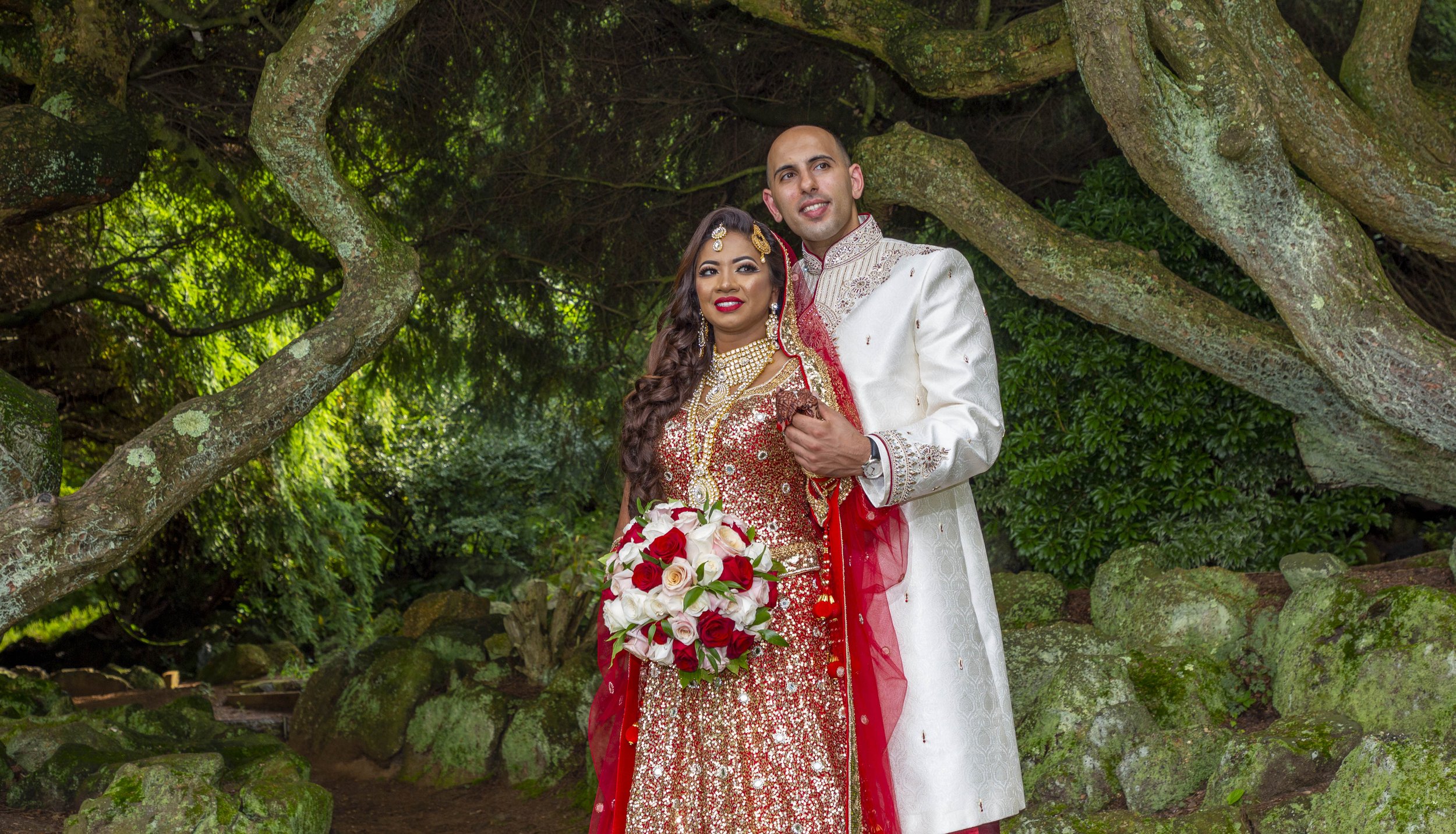 A couple in traditional South Asian wedding attire pose under a large tree. The woman wears a red and gold embellished dress with ornate jewelry and holds a bouquet of red and white roses. The man wears a white embroidered sherwani. The background fe