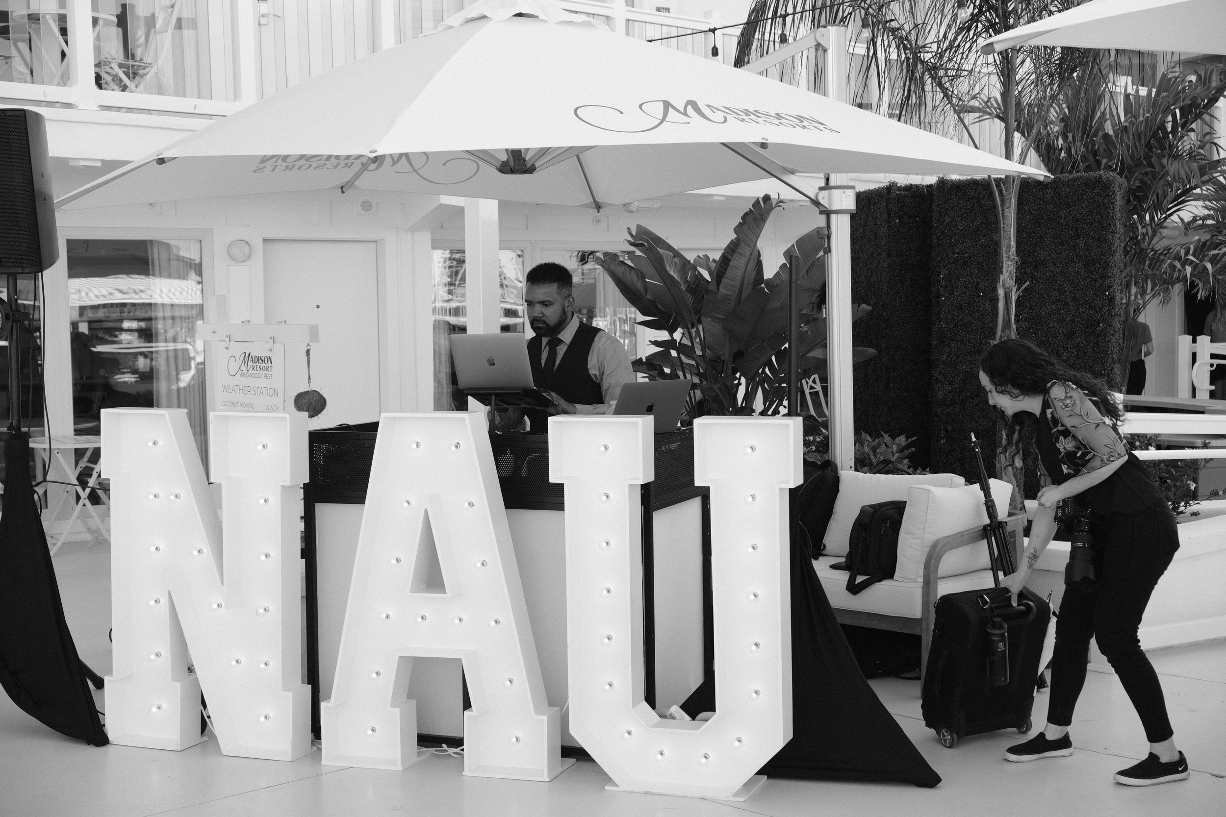 DJ in an outdoor setting with NAU marquee letters, woman arranging equipment, large umbrella, modern decor.