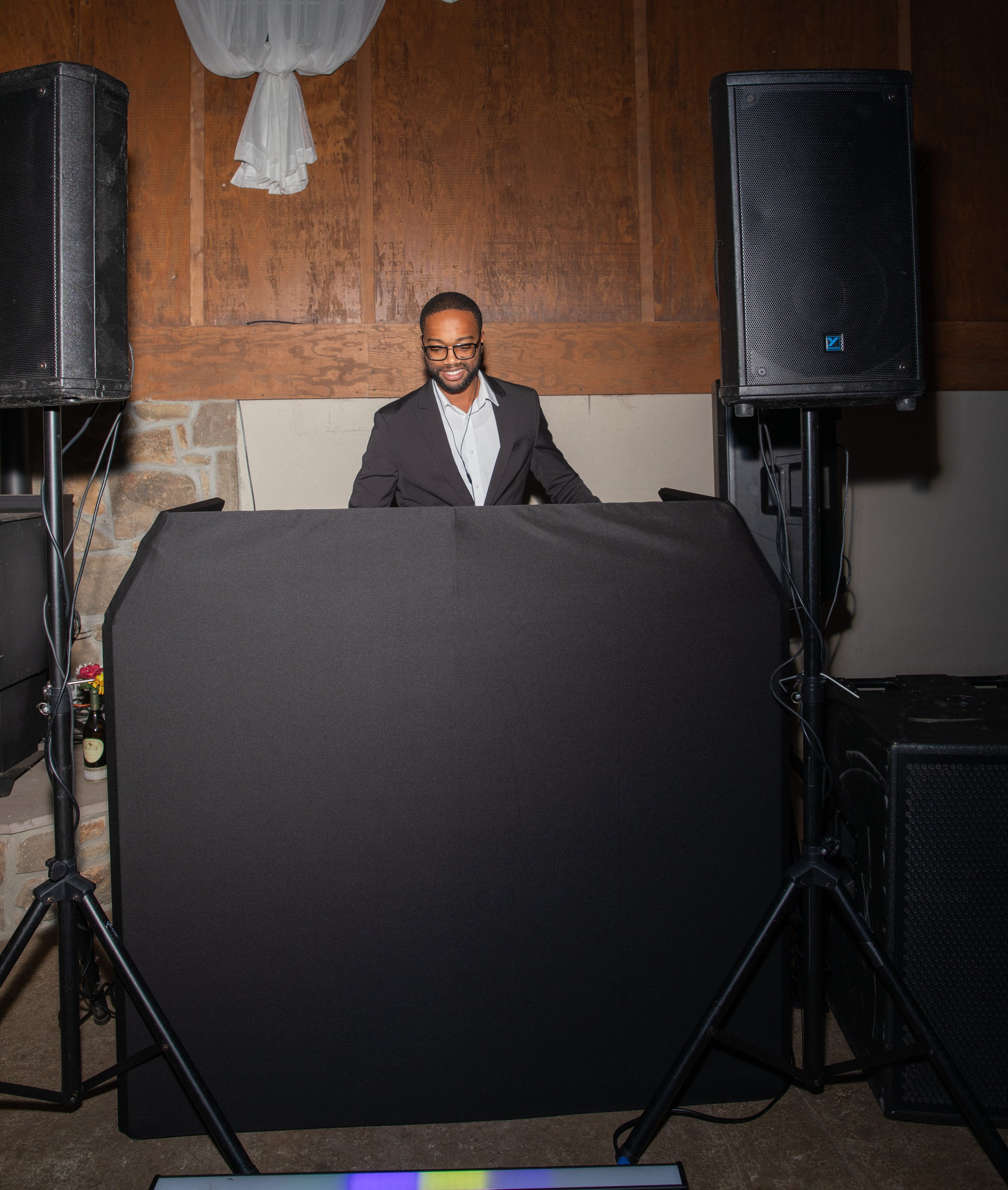 DJ stands behind a covered table with speakers and equipment at an event.