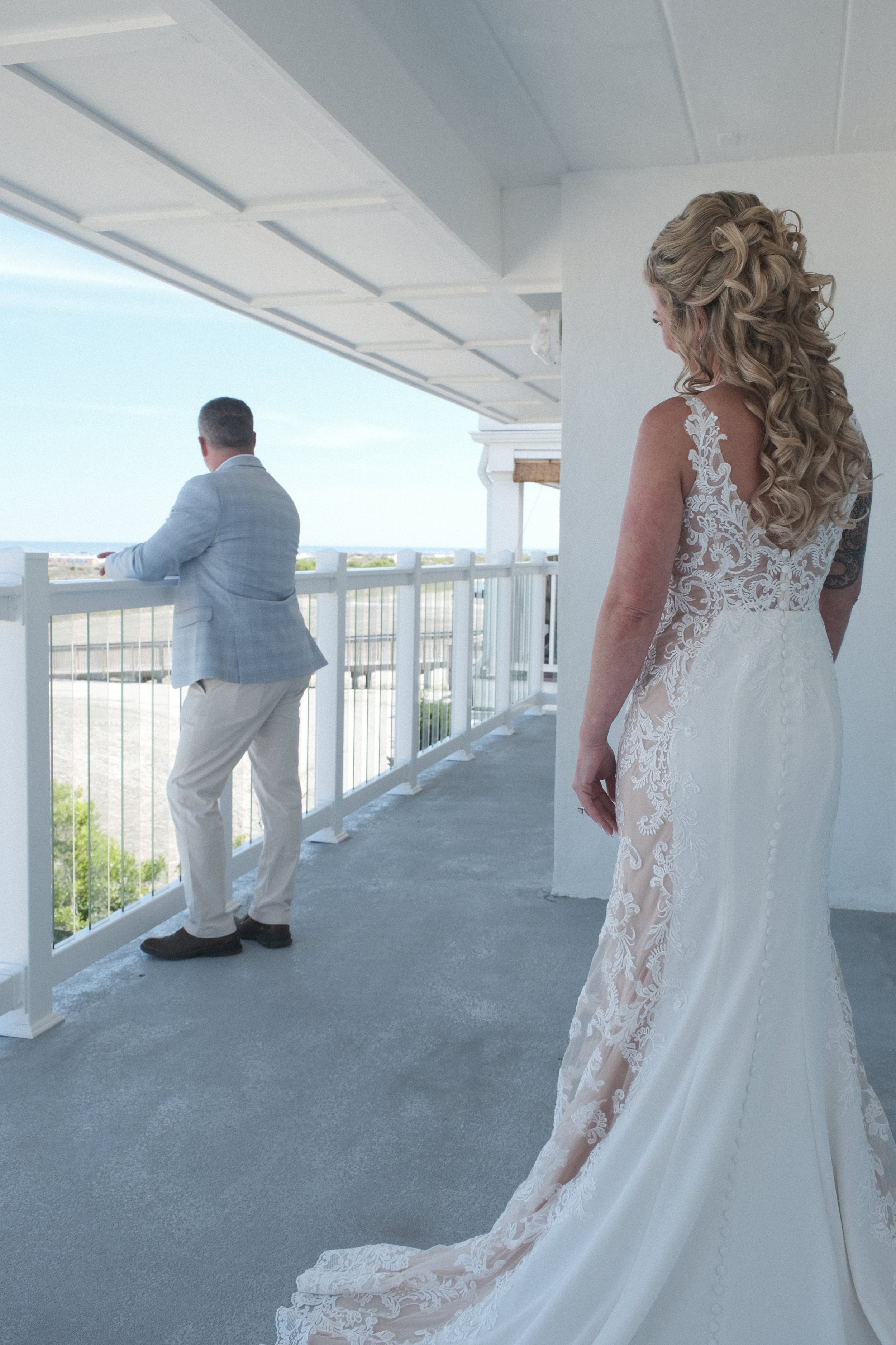 Bride in lace wedding gown approaching groom on a balcony overlooking a scenic view.