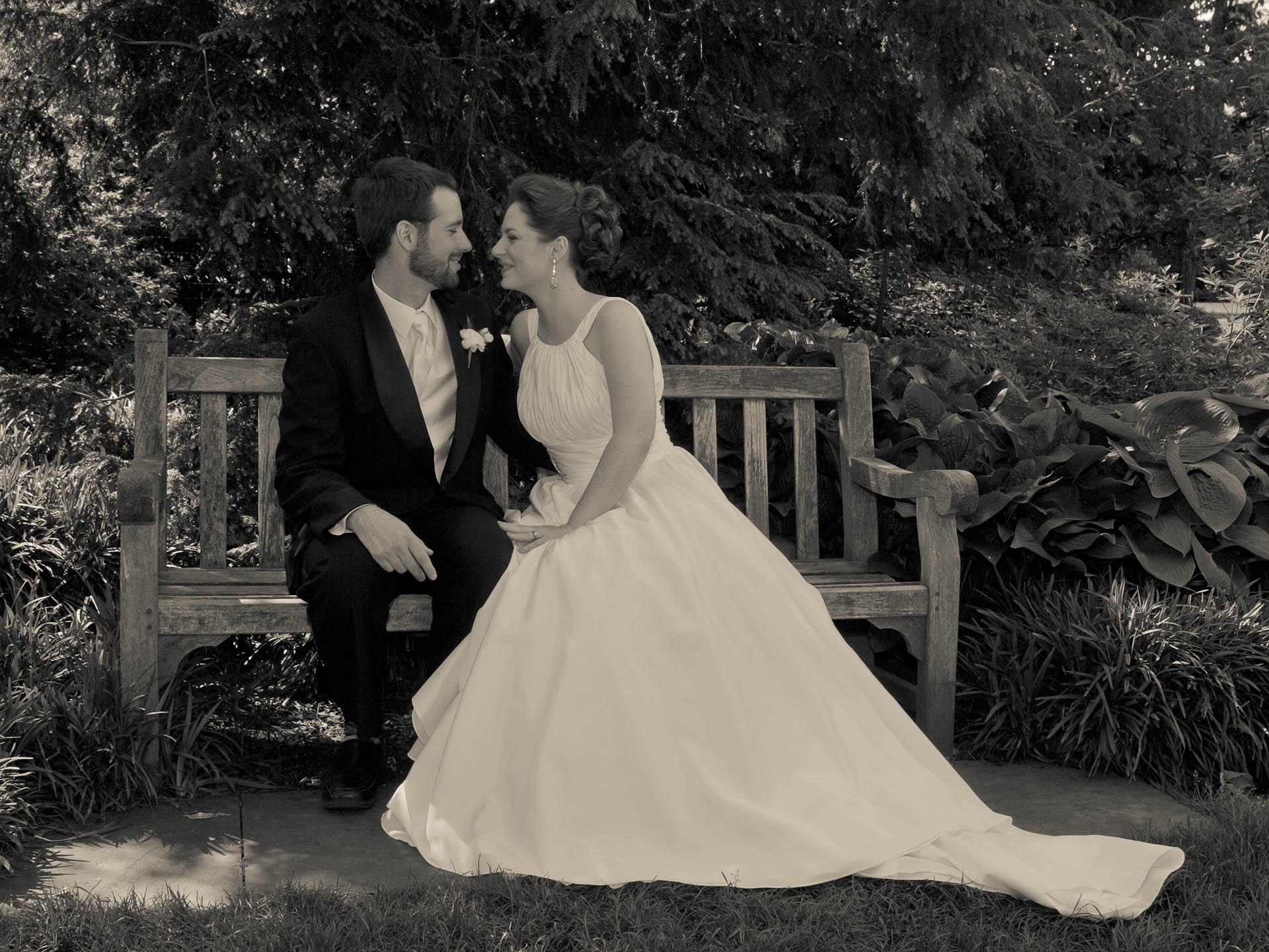Bride and groom sitting on a bench in a garden setting, dressed in wedding attire, with trees and foliage in the background.
