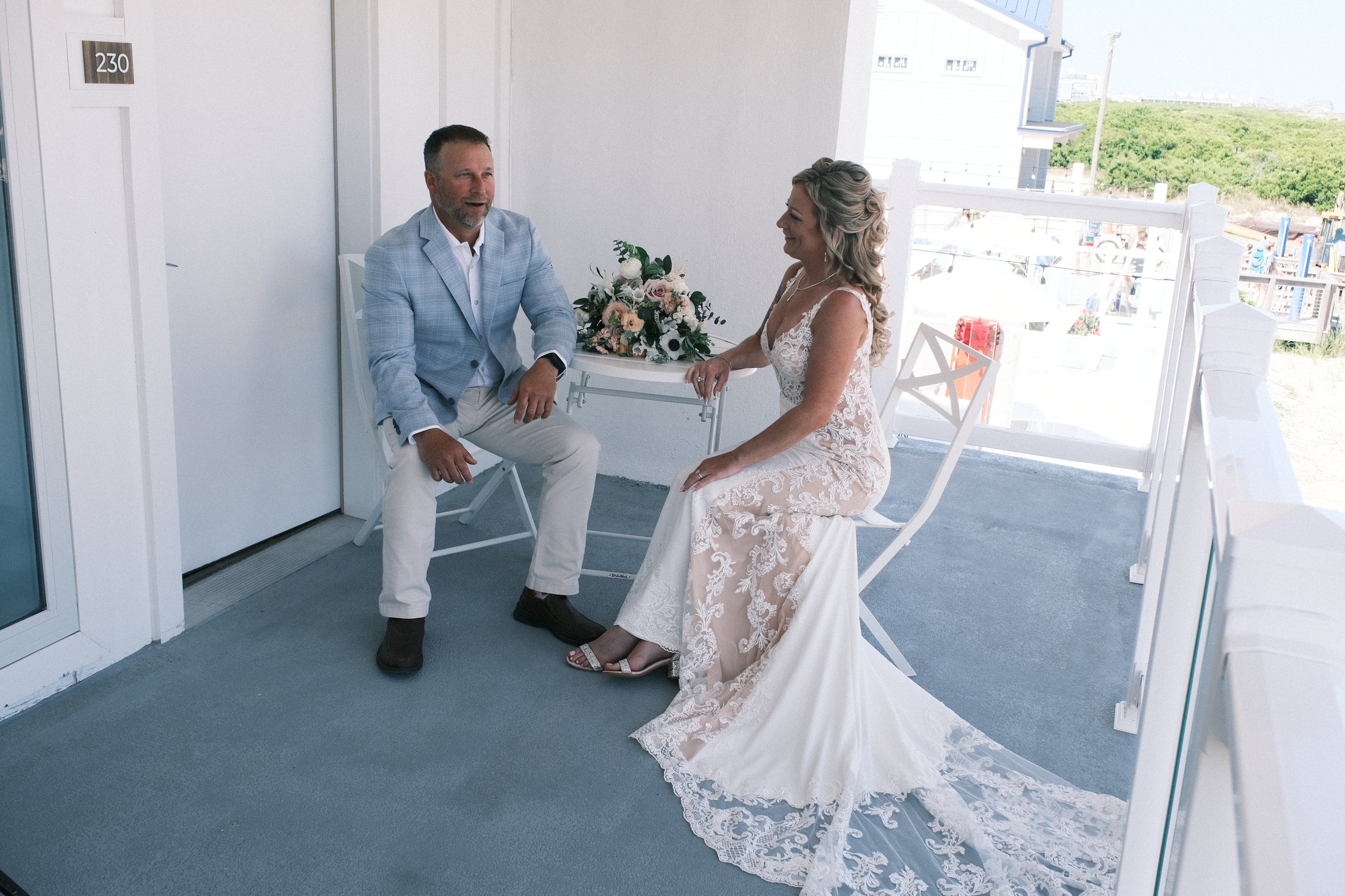 A man in a light blue blazer and beige pants sits across from a woman in a lace wedding dress on a balcony with white decor and a bouquet on the table.