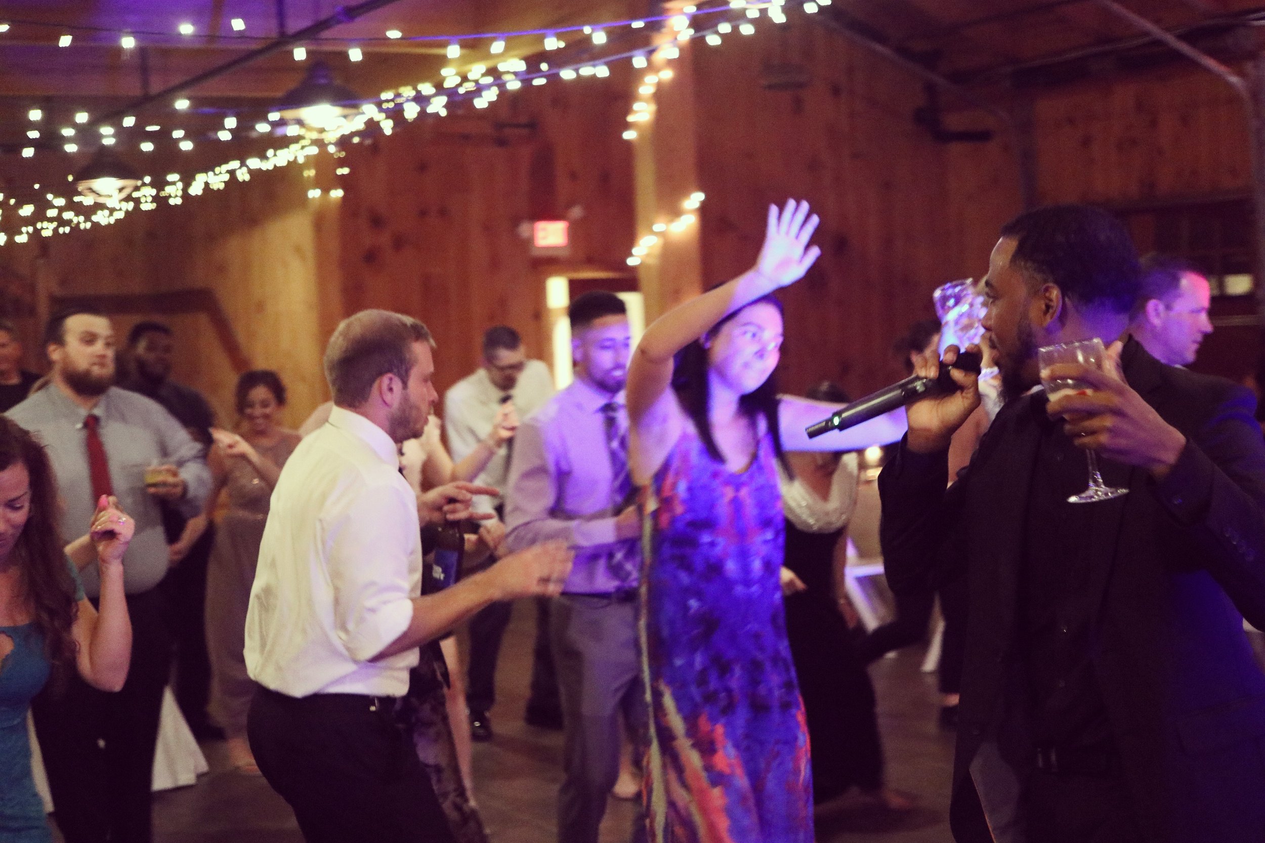 People dancing and celebrating at an indoor party under string lights, with a man holding a microphone and a glass of wine.