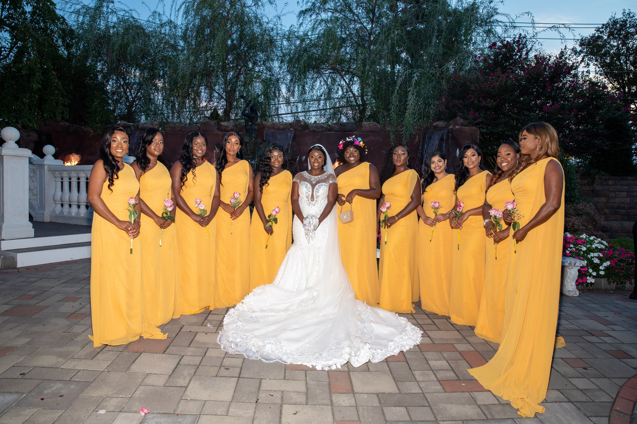 Bride in white gown surrounded by bridesmaids in yellow dresses, outdoor setting with greenery.