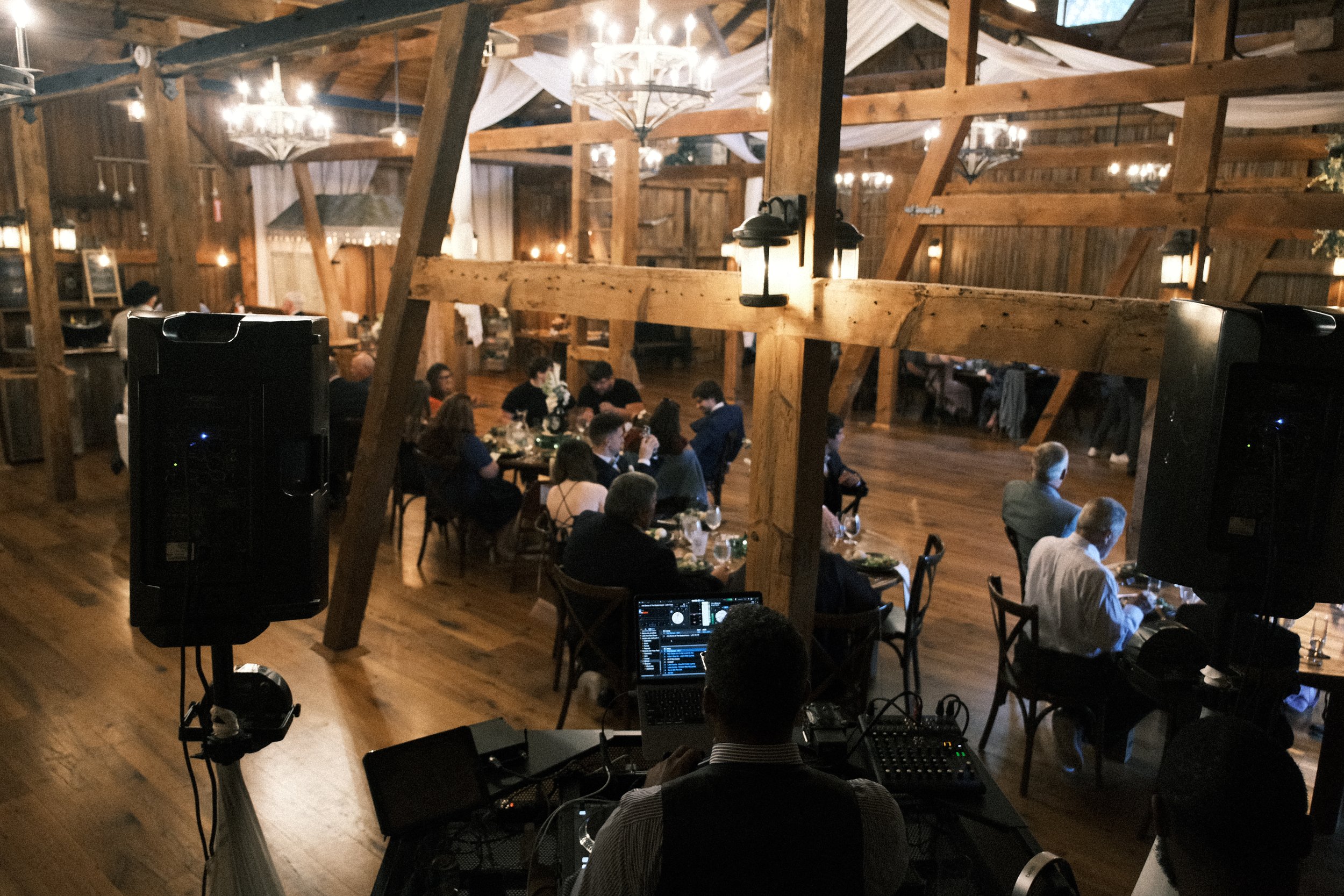 DJ at a rustic event venue with wooden beams, playing music for a seated audience at round tables under chandeliers.