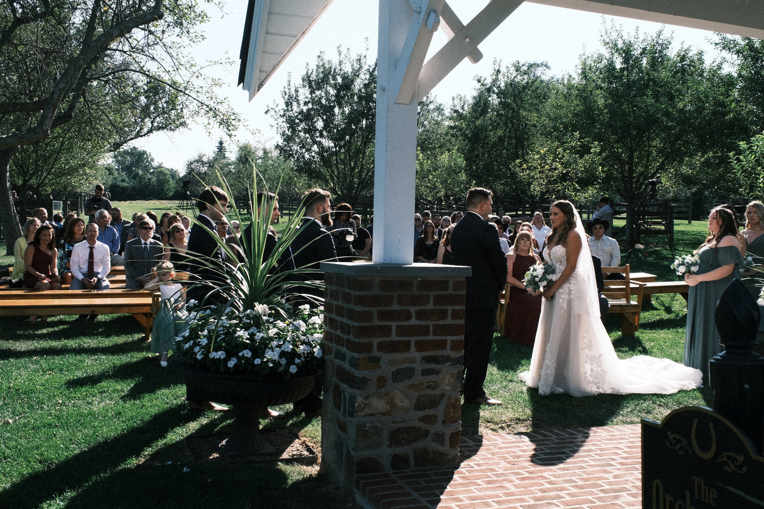 Outdoor wedding ceremony with bride and groom, guests seated on wooden benches, surrounded by green trees, sunlight, and floral decorations.